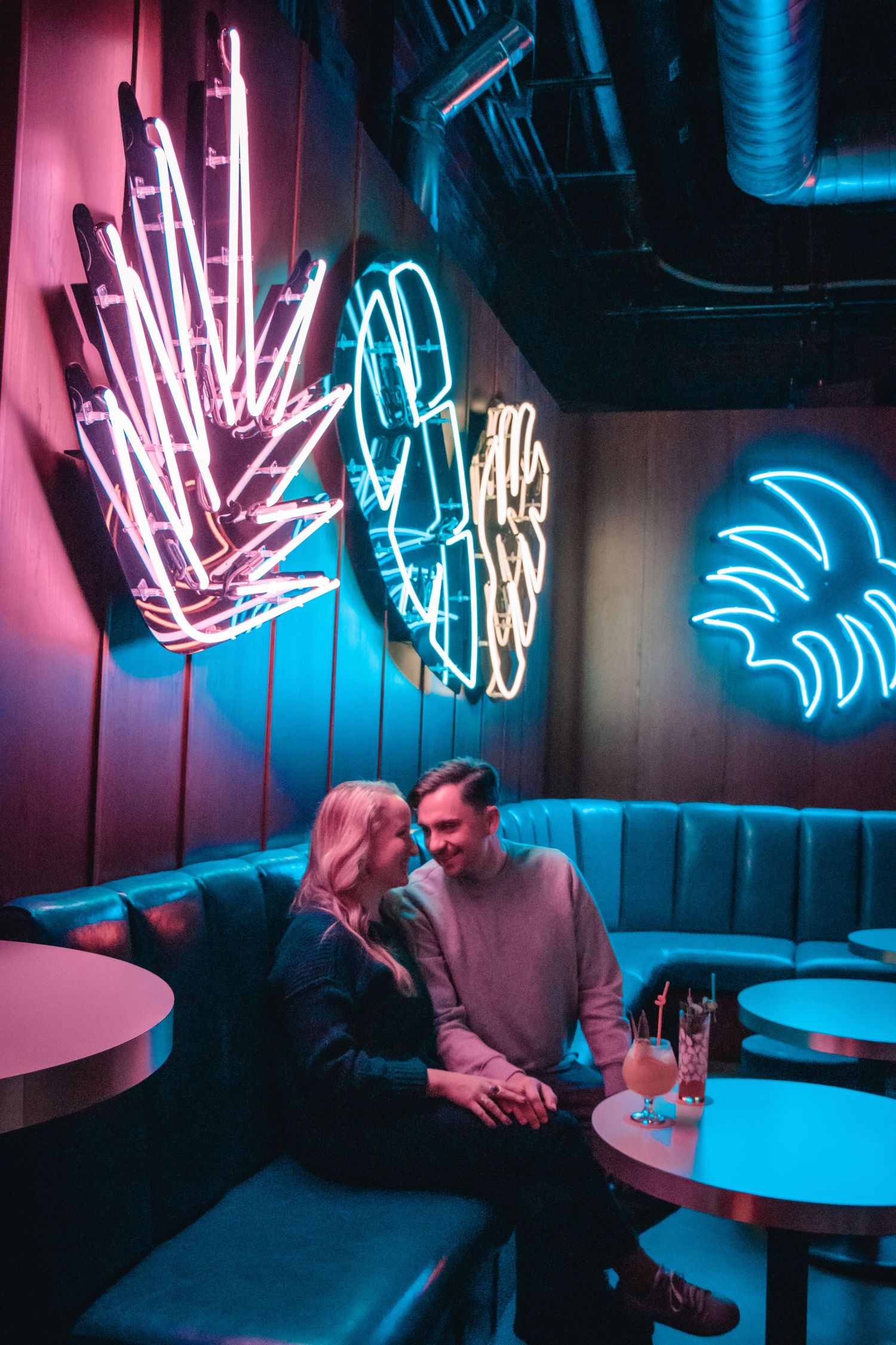 A couple sits in a retro bar booth illuminated by neon palm tree signs on the wall.