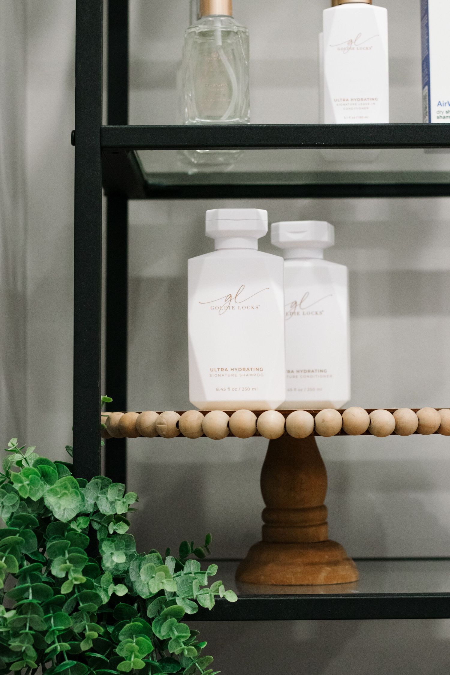 White bottles displayed on wooden beaded cake stand with greenery on black shelf.