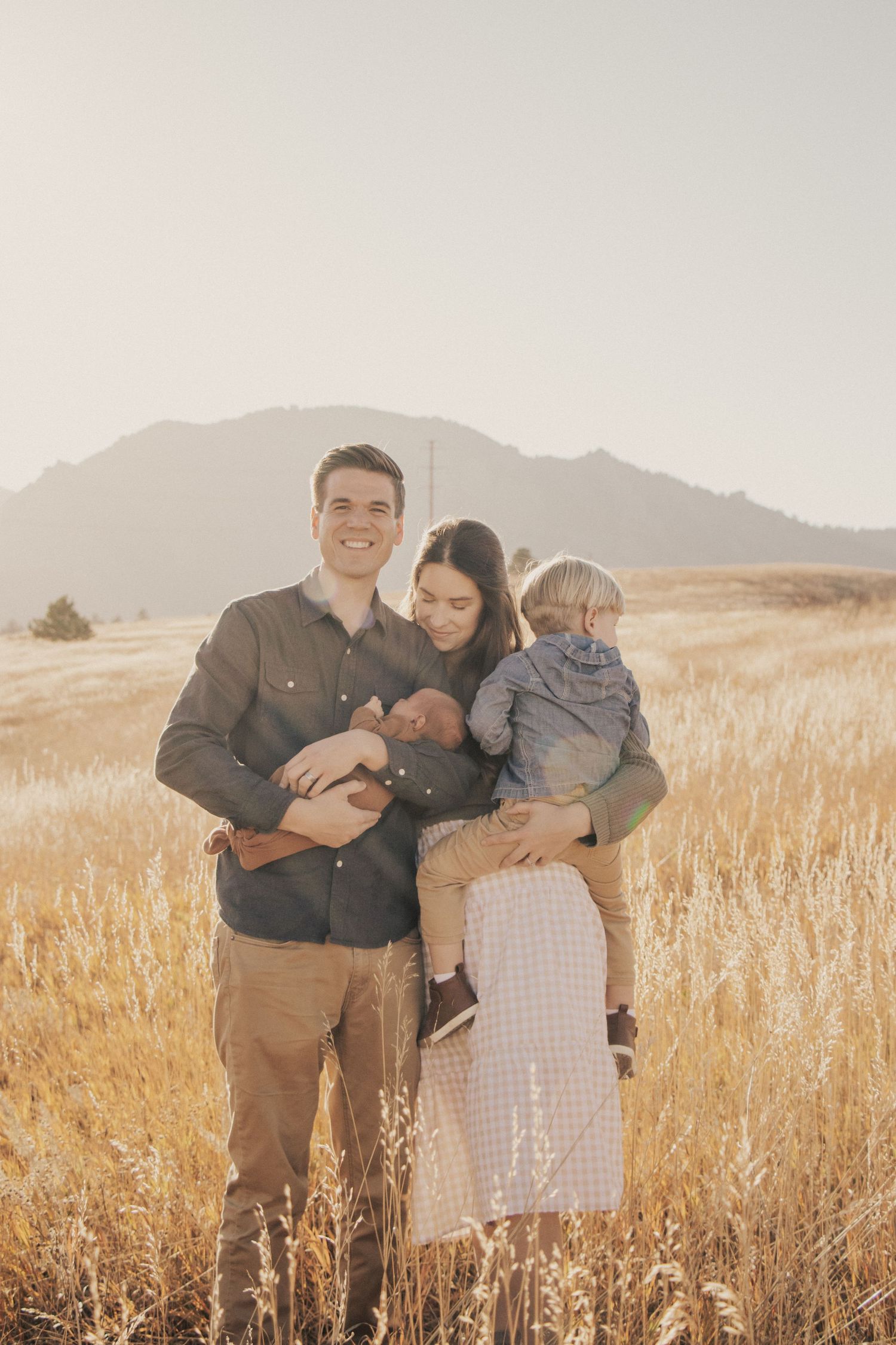 Family of four poses together in sun-drenched field with mountain backdrop.