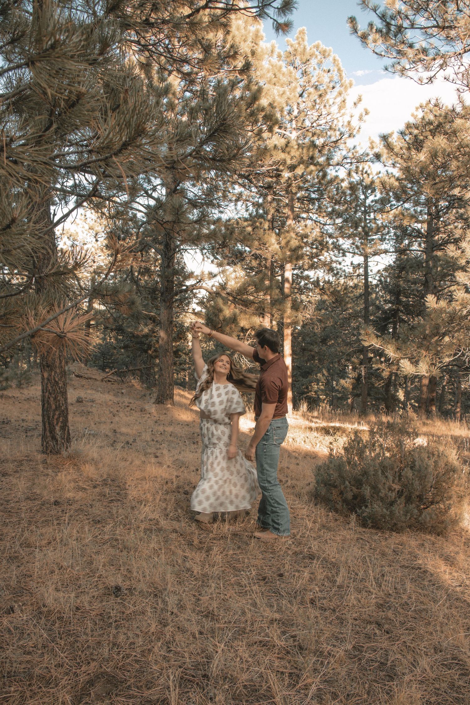 Couple standing together on pine-covered hillside in warm afternoon light.