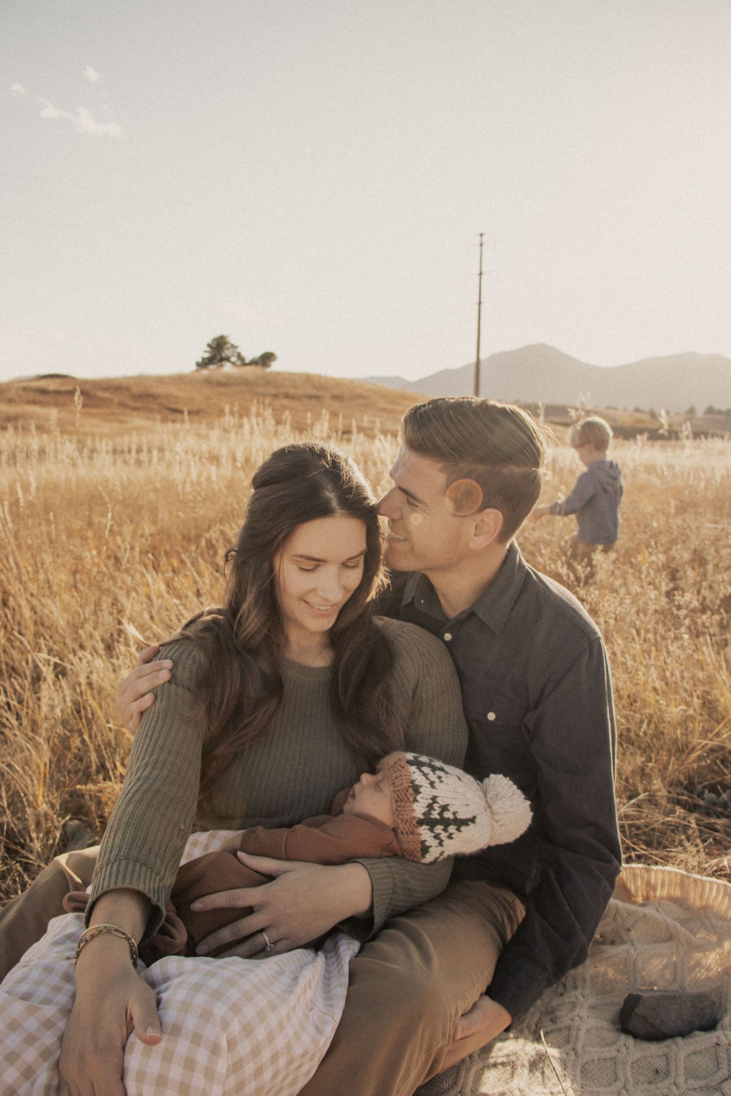 A couple sits in a golden field at sunset cradling their newborn baby with mountains in the background.