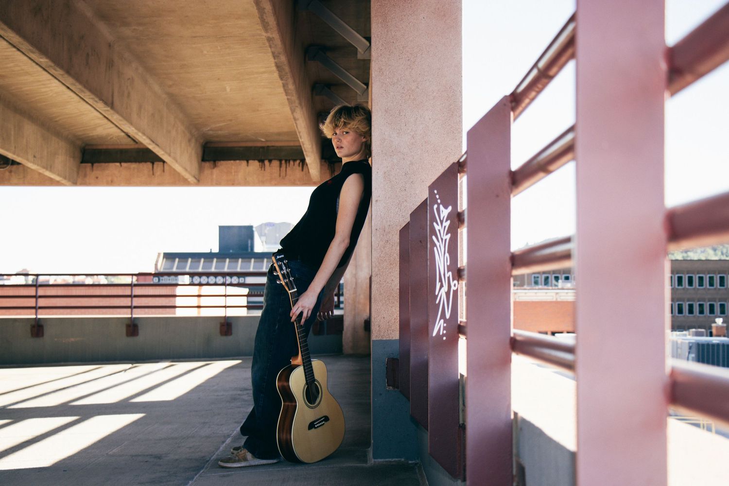 A musician stands with guitar beside a concrete pillar in an urban setting.