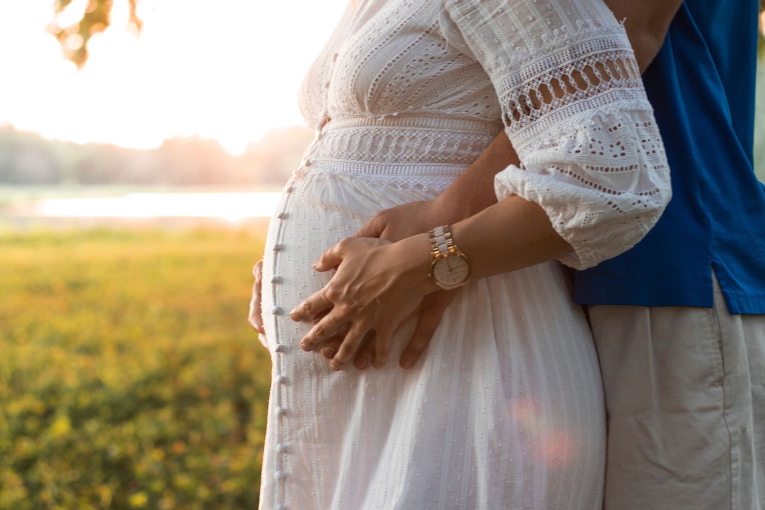 Close up of hands cradling pregnant belly in white lace dress at sunset.
