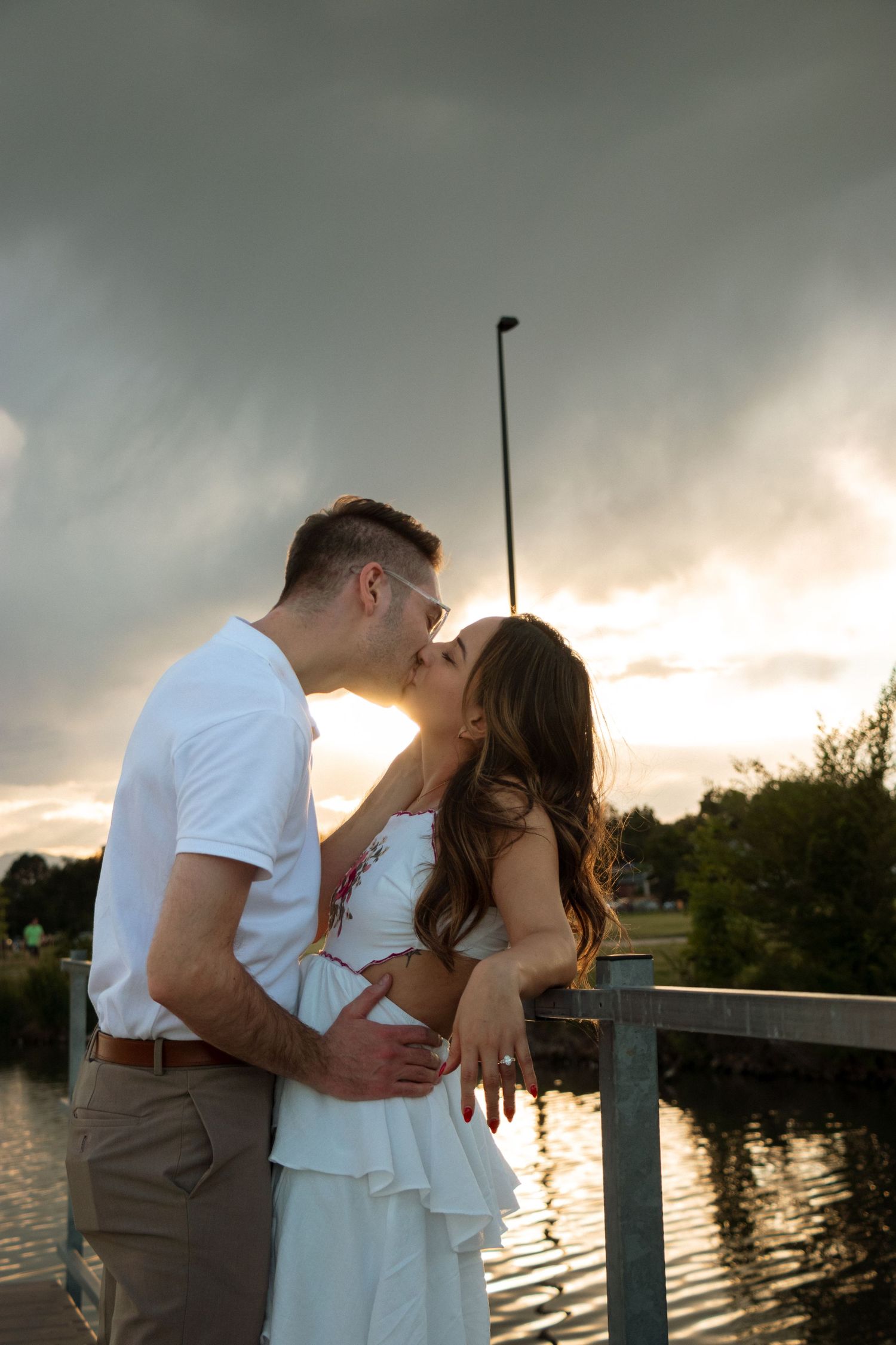 A romantic couple shares intimate moments at sunset by a waterfront dock with dramatic cloudy skies in the background.