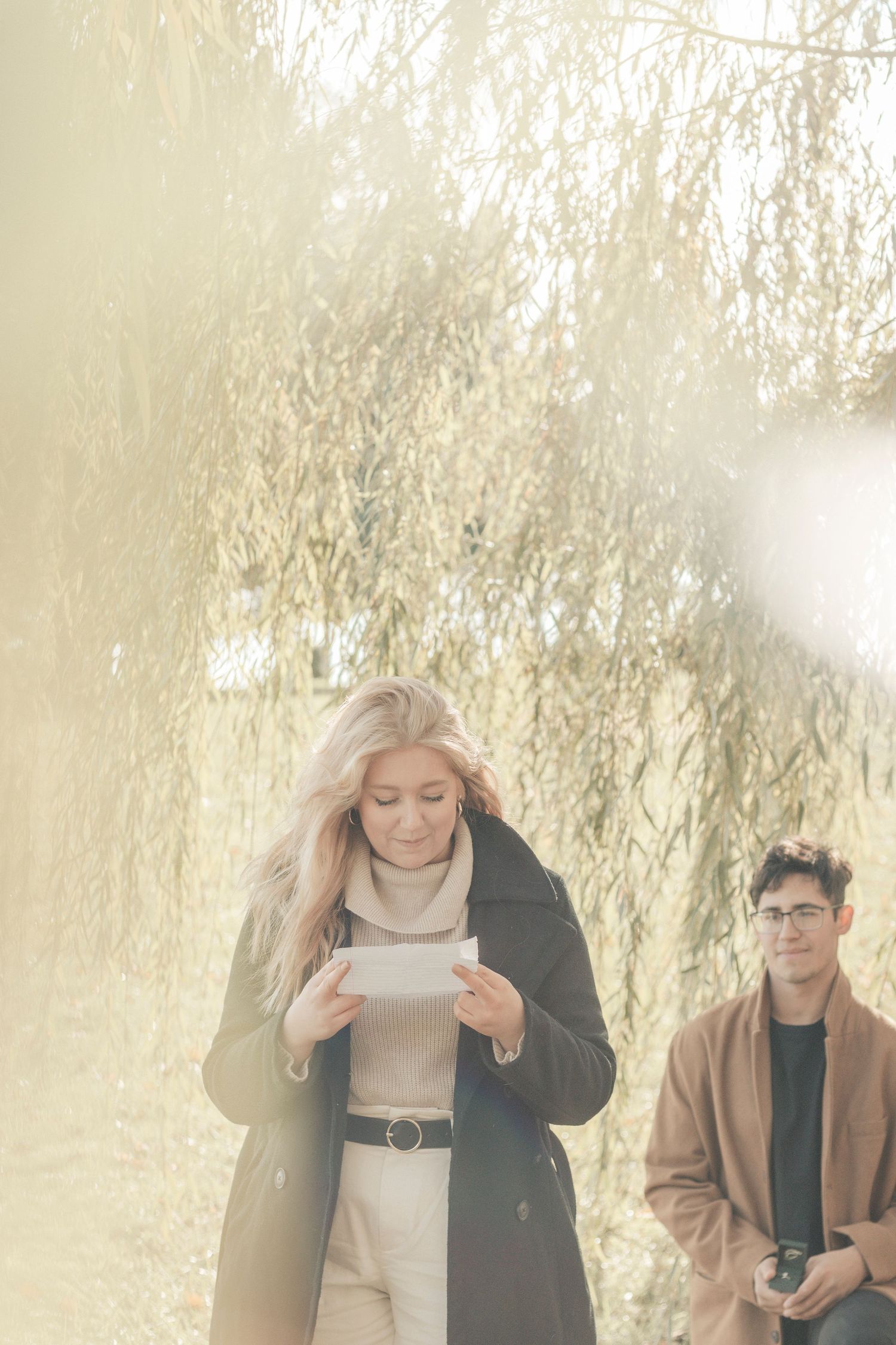 Two people in winter coats standing beneath willow tree branches in soft sunlight.