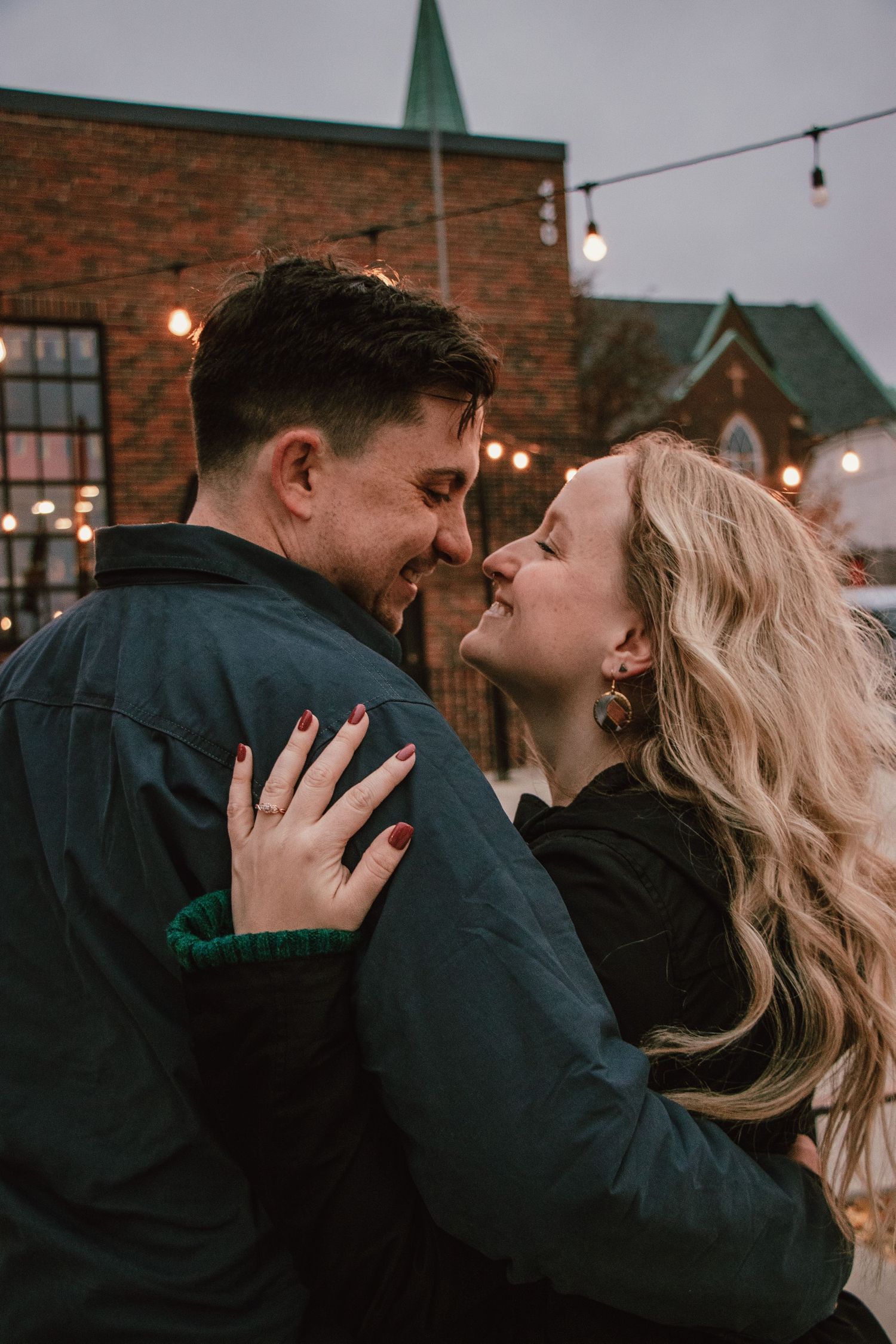 A romantic couple shares an intimate moment beneath string lights with a church spire visible in the background.