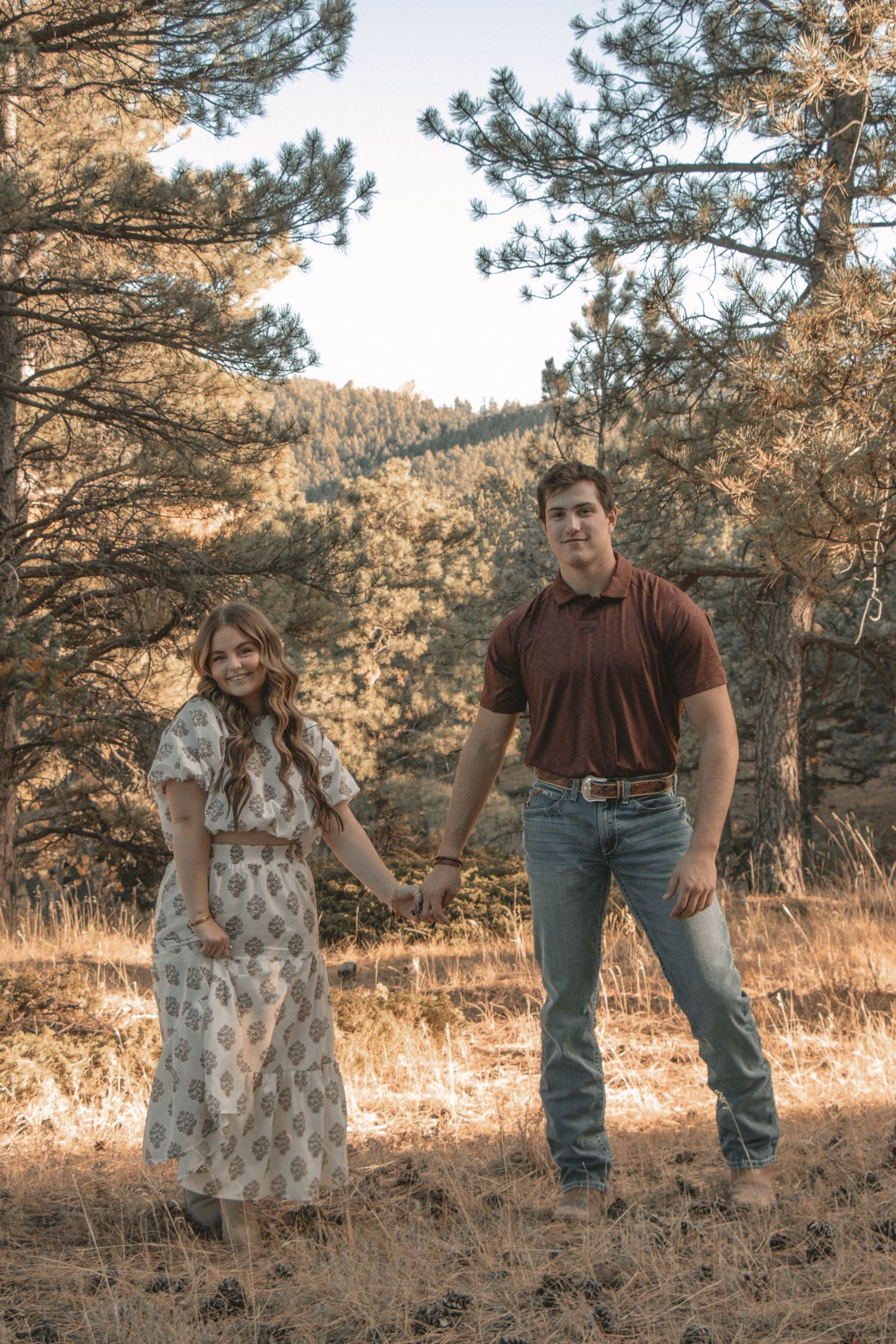 Couple in casual attire posing together on wooded hillside.