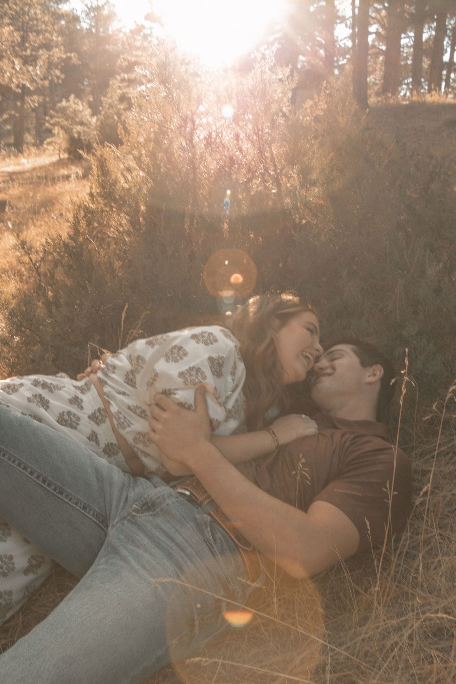 Couple lying in tall grass under pine trees at golden hour.