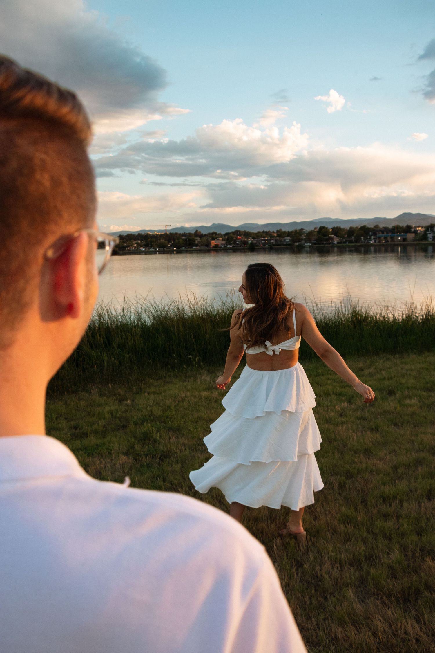 Couple stands near a lakeside at sunset as woman in white dress runs through tall grass with mountains in background.