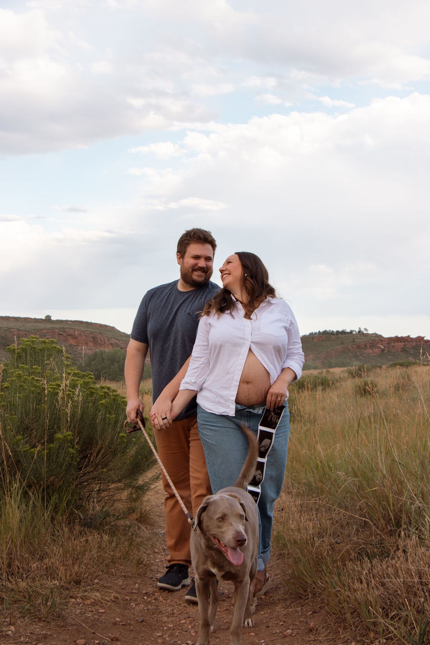 A couple and their dog enjoy a hike together on a scenic trail with hills and clouds in the background.