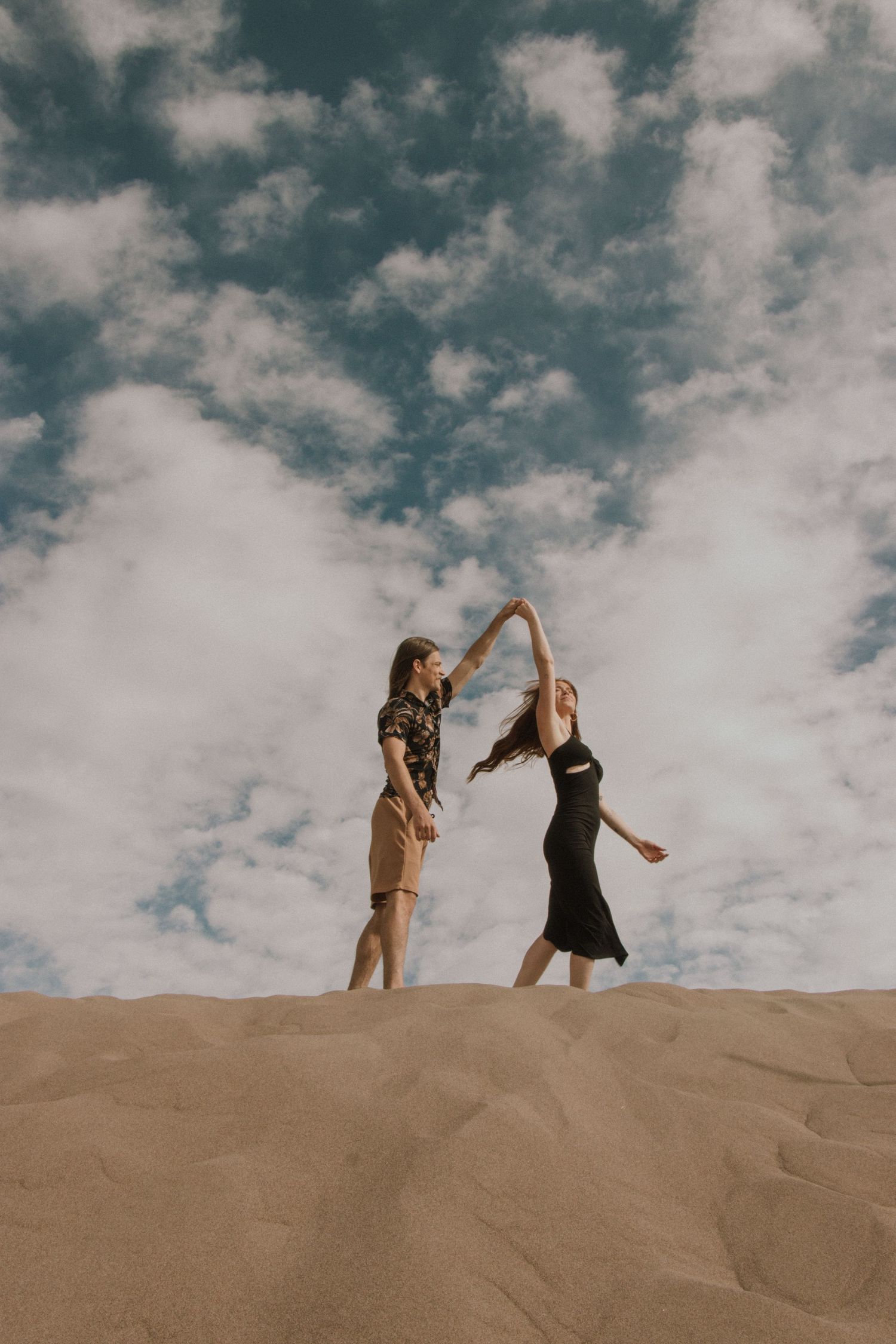 Two people dancing and twirling on sand dunes under a dramatic cloudy sky.