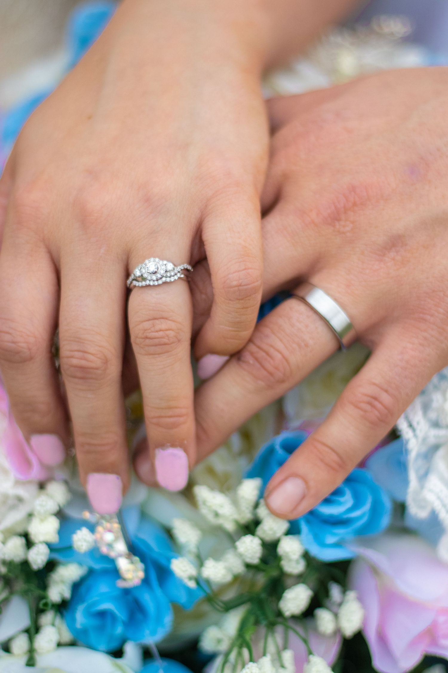 Close-up of newlyweds' hands showing wedding rings against blue and pink floral bouquet.