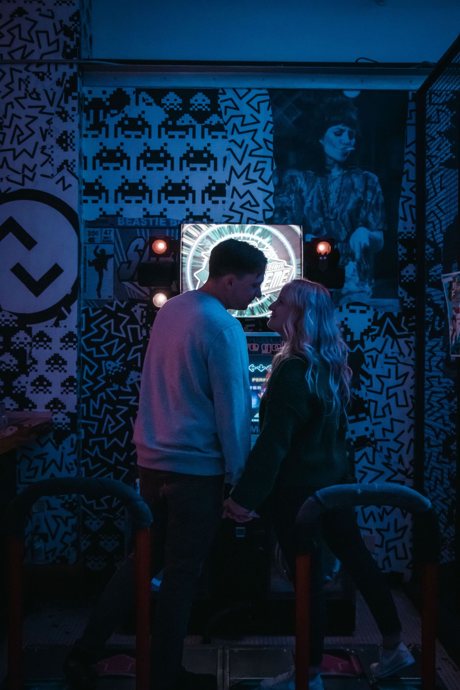 Two people playing arcade games against a backdrop of retro pixel art wallpaper.