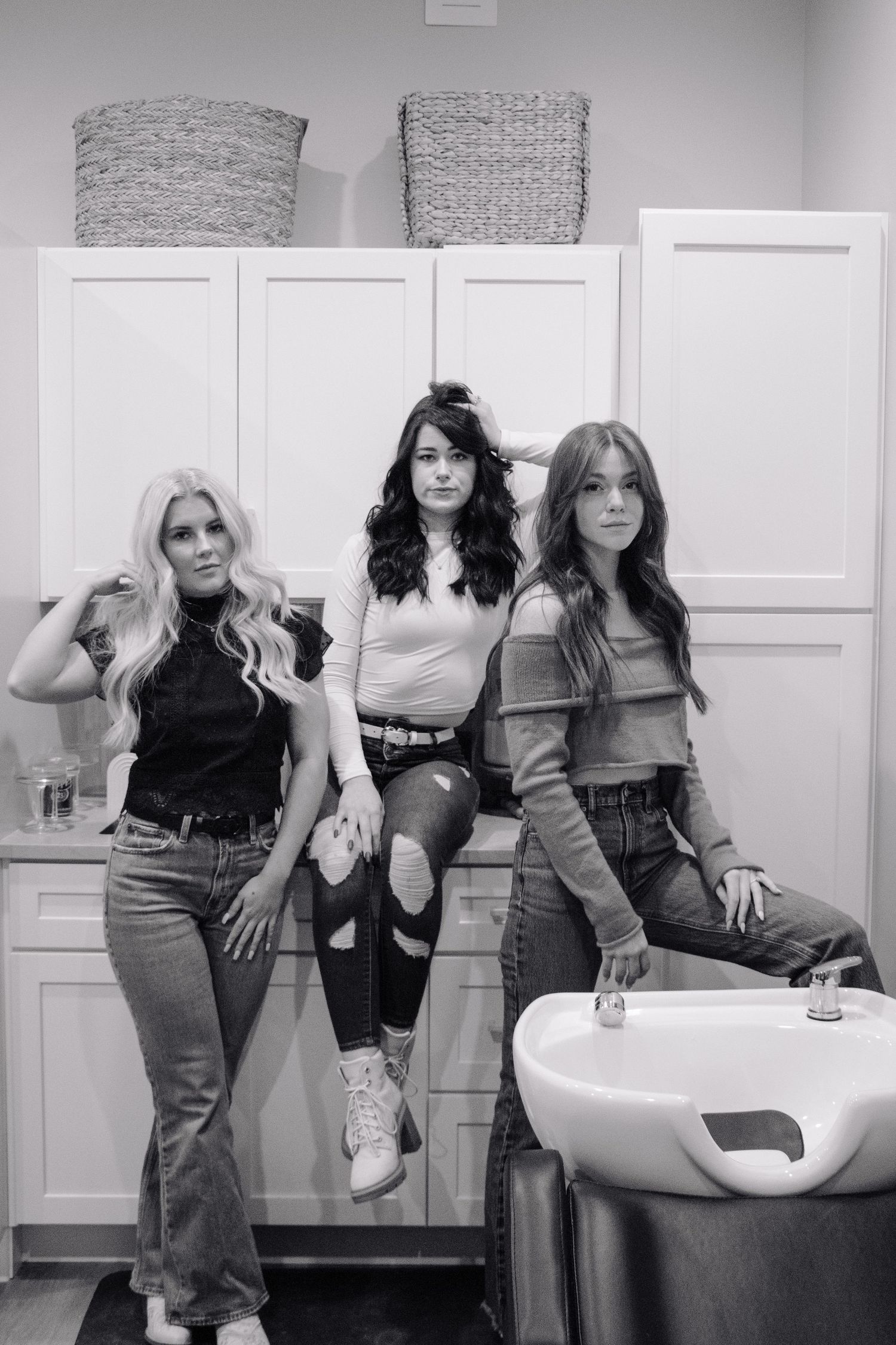 Three people in casual retro-style outfits pose in a salon setting with white cabinets and a hair washing station.
