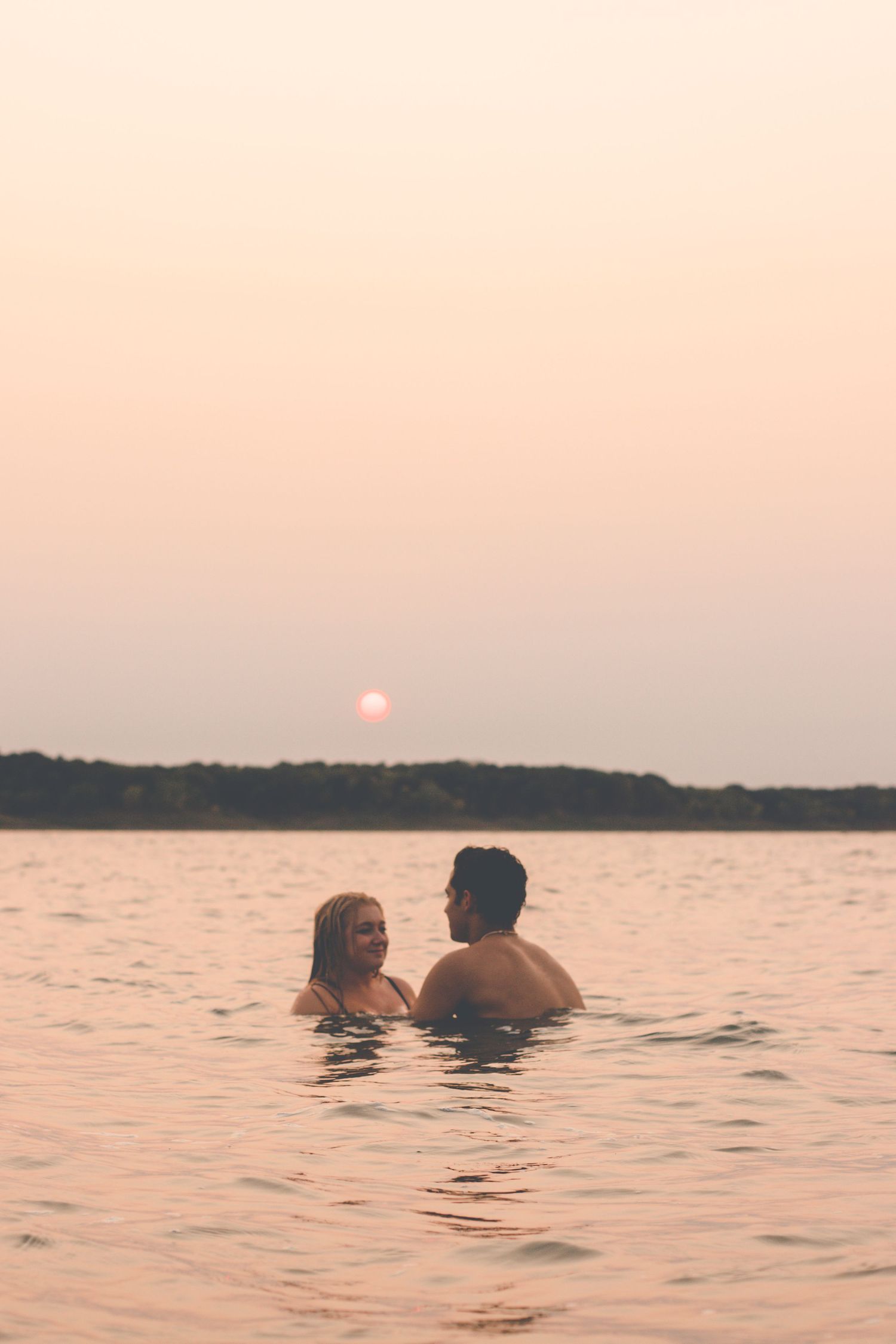 Couple swimming together in lake waters during pink sunset.