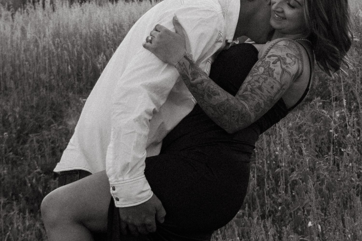 Black and white photo of a tattooed couple embracing romantically in a field of tall grass.
