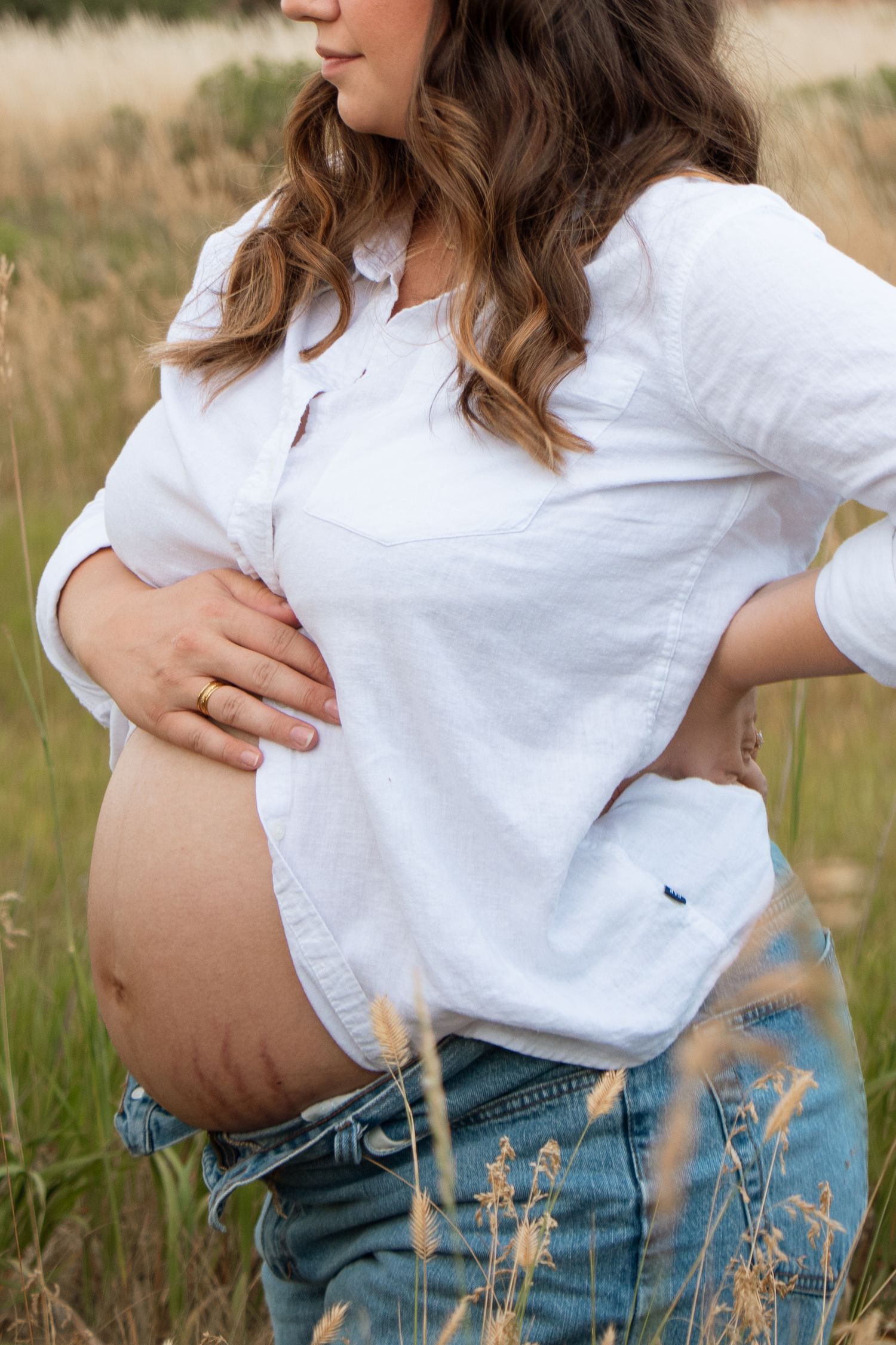 A maternity photo series showing someone wearing white top and denim in a golden grass field during sunset.