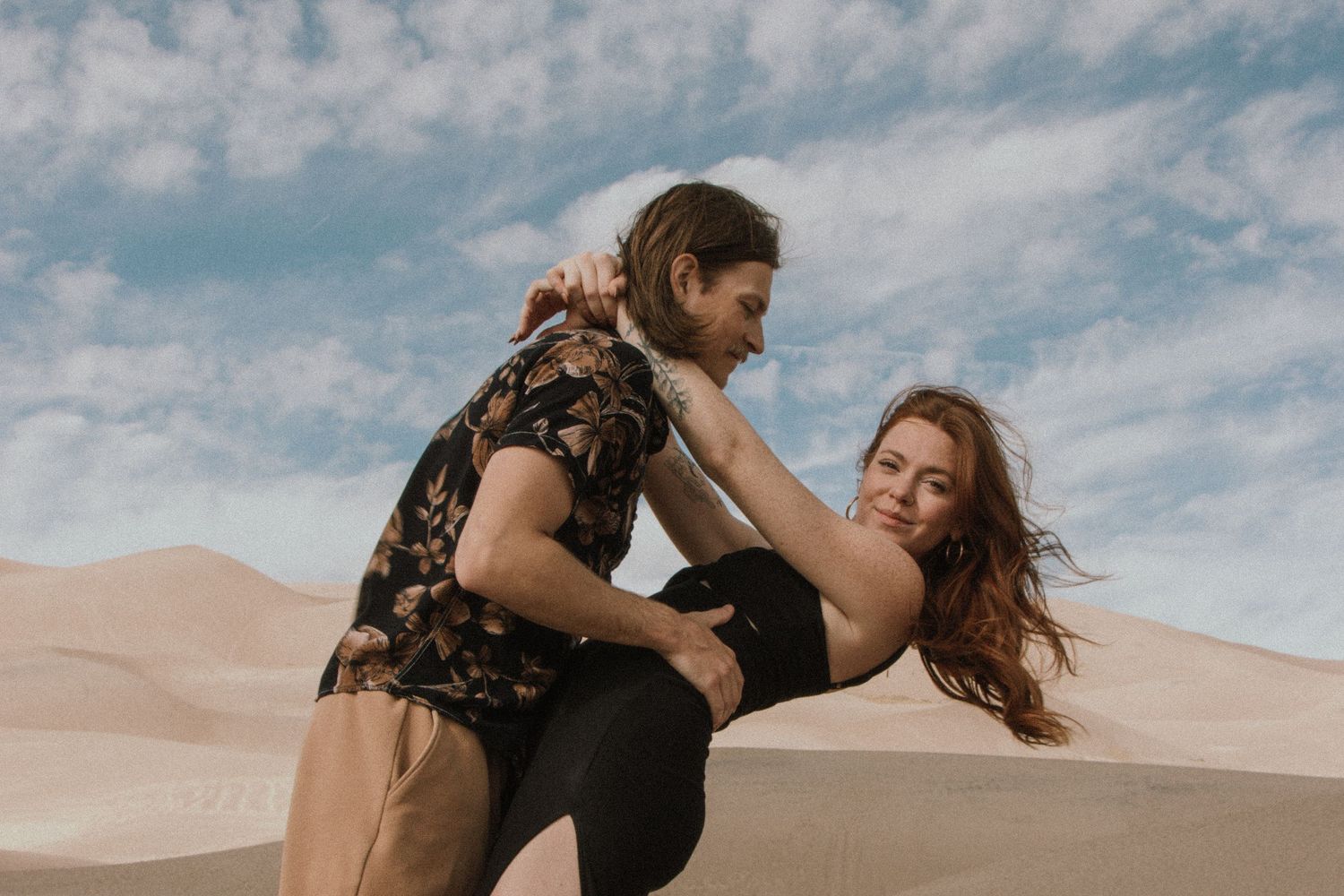 A playful moment between two people dancing in sand dunes against a cloudy blue sky.