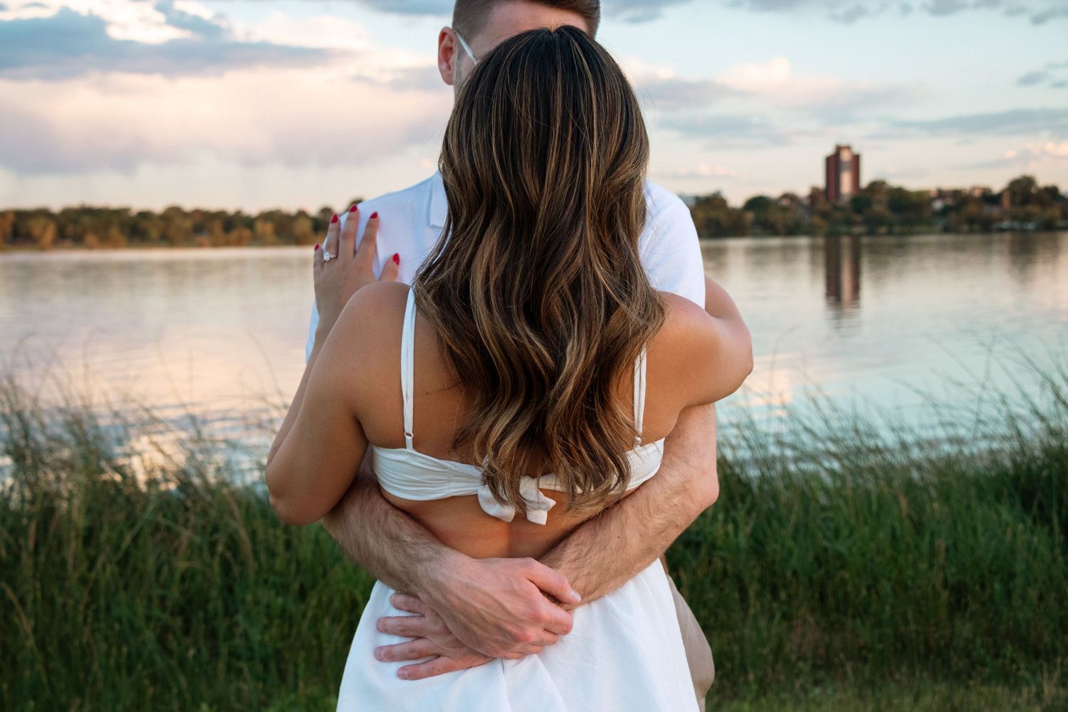 Couple embraces on a dock by a tranquil lake at sunset with tall grass and clouds in the background.