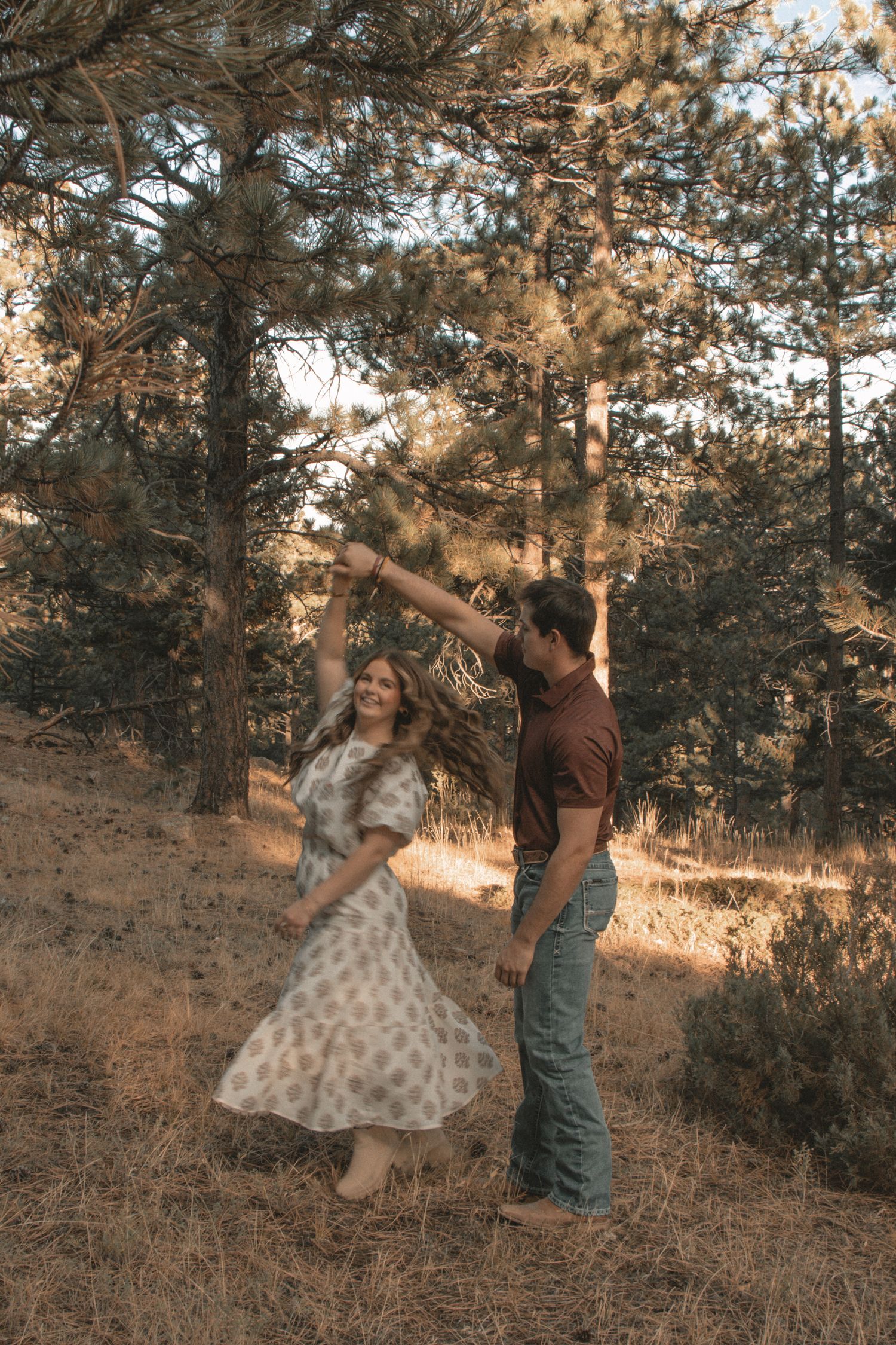 Couple dancing and twirling in vintage clothing among pine trees at sunset.
