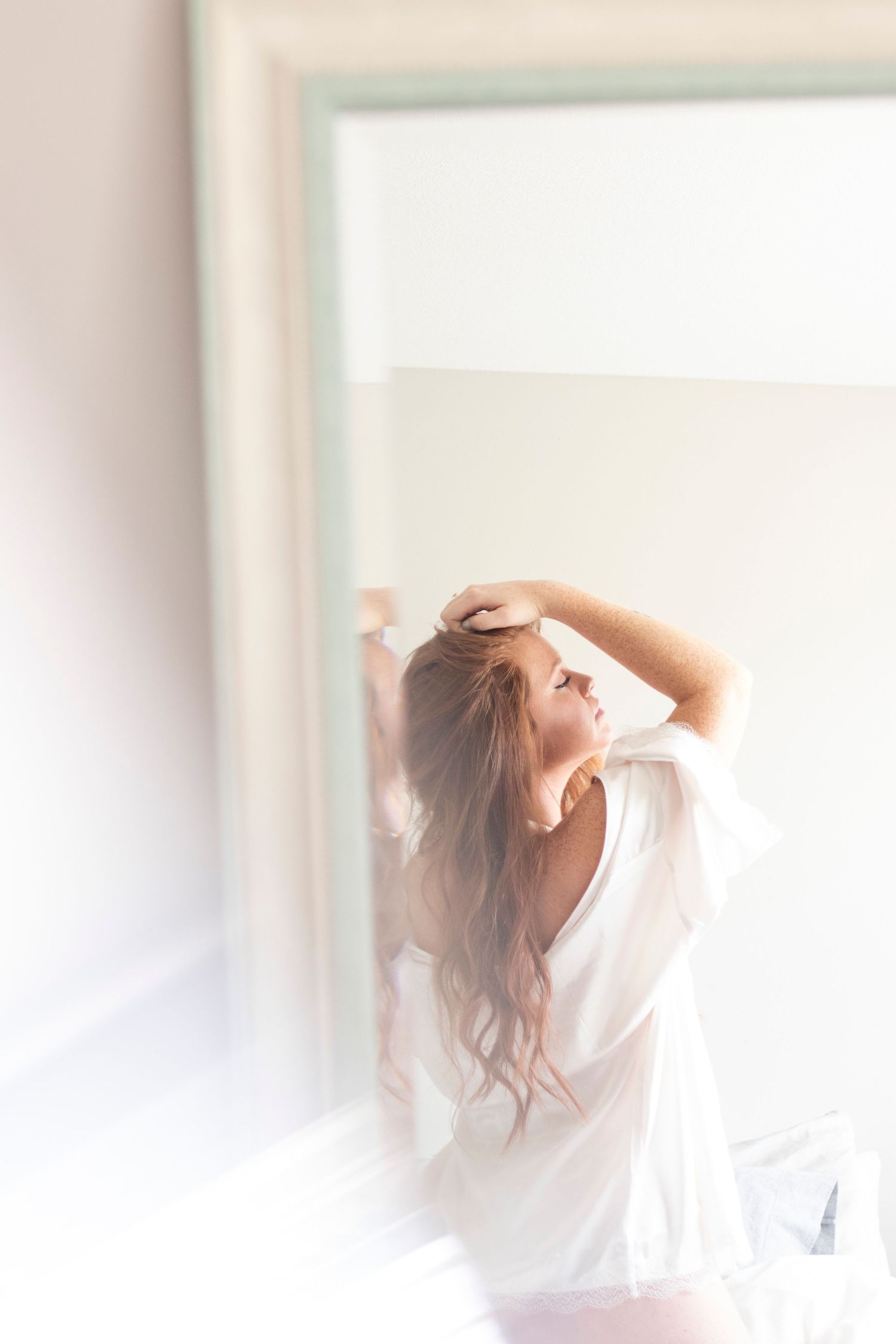 Soft dreamy portrait of woman stretching in white flowing dress by window.