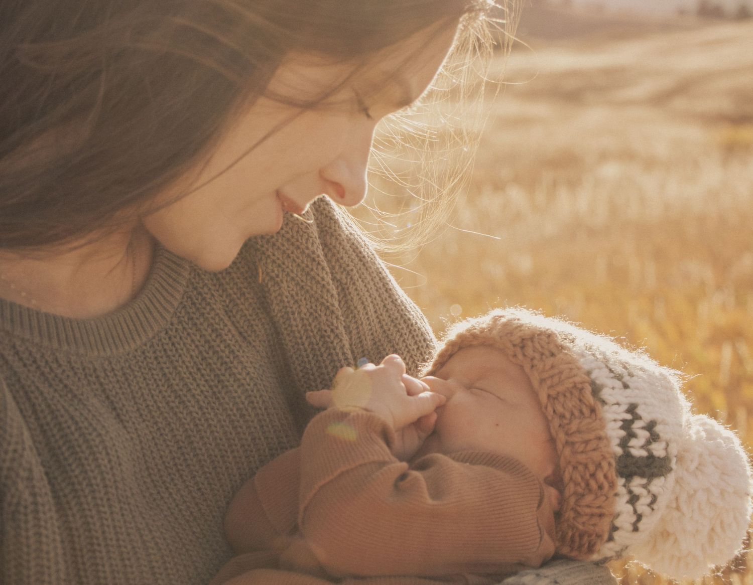 Parent cuddles with newborn baby wearing knit hat in warm sunlight.