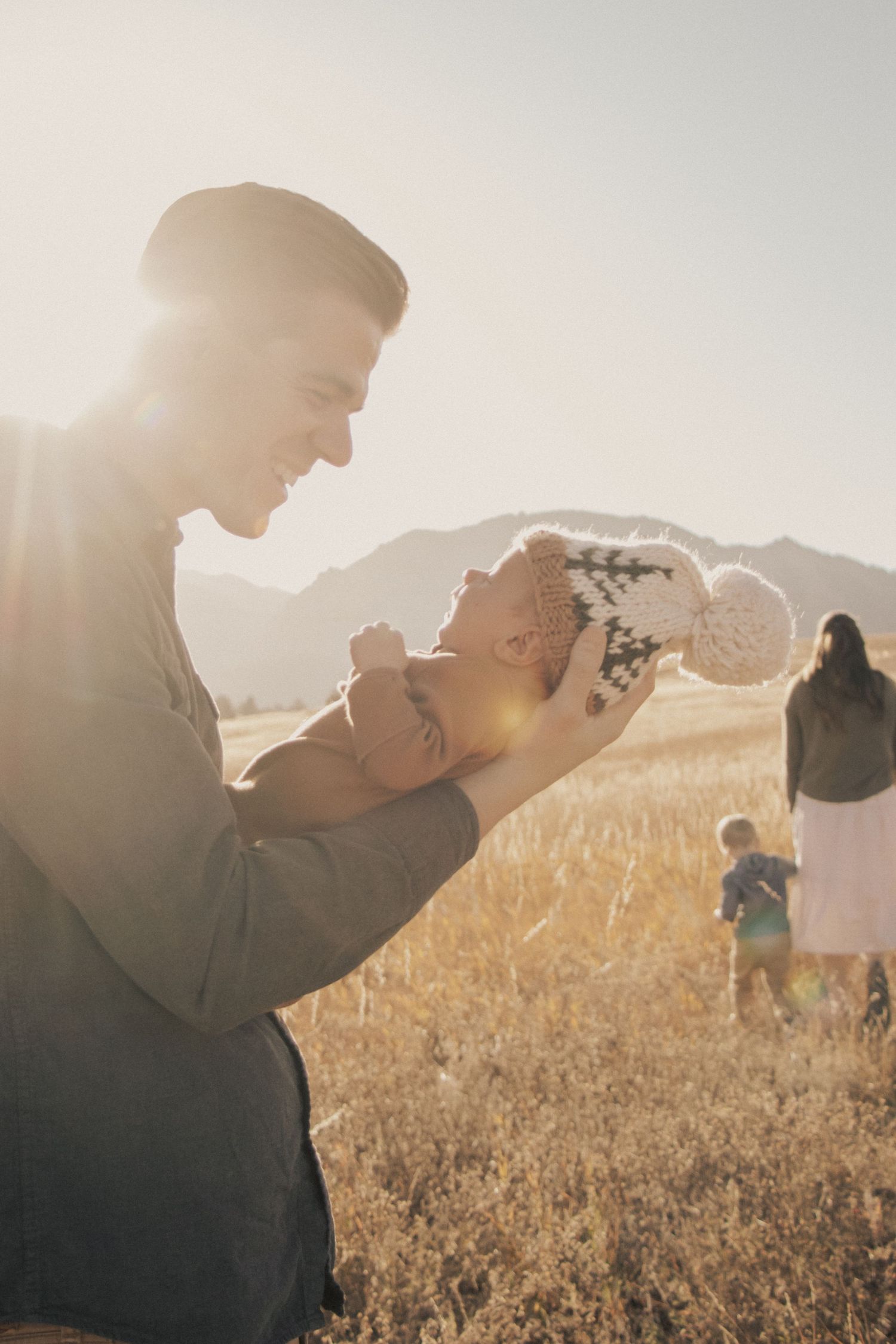 Parent holds up small infant in sunlit field with mountains and family members visible in background.