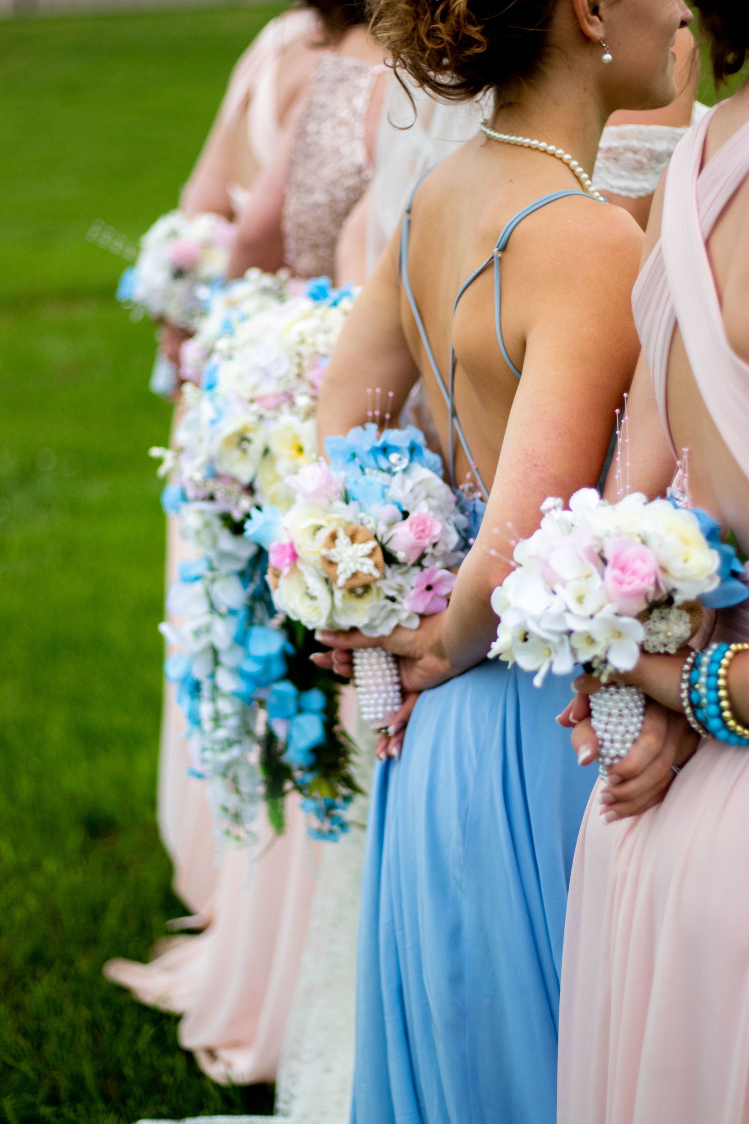Wedding party members hold pastel bouquets featuring blue and pink flowers against green grass.