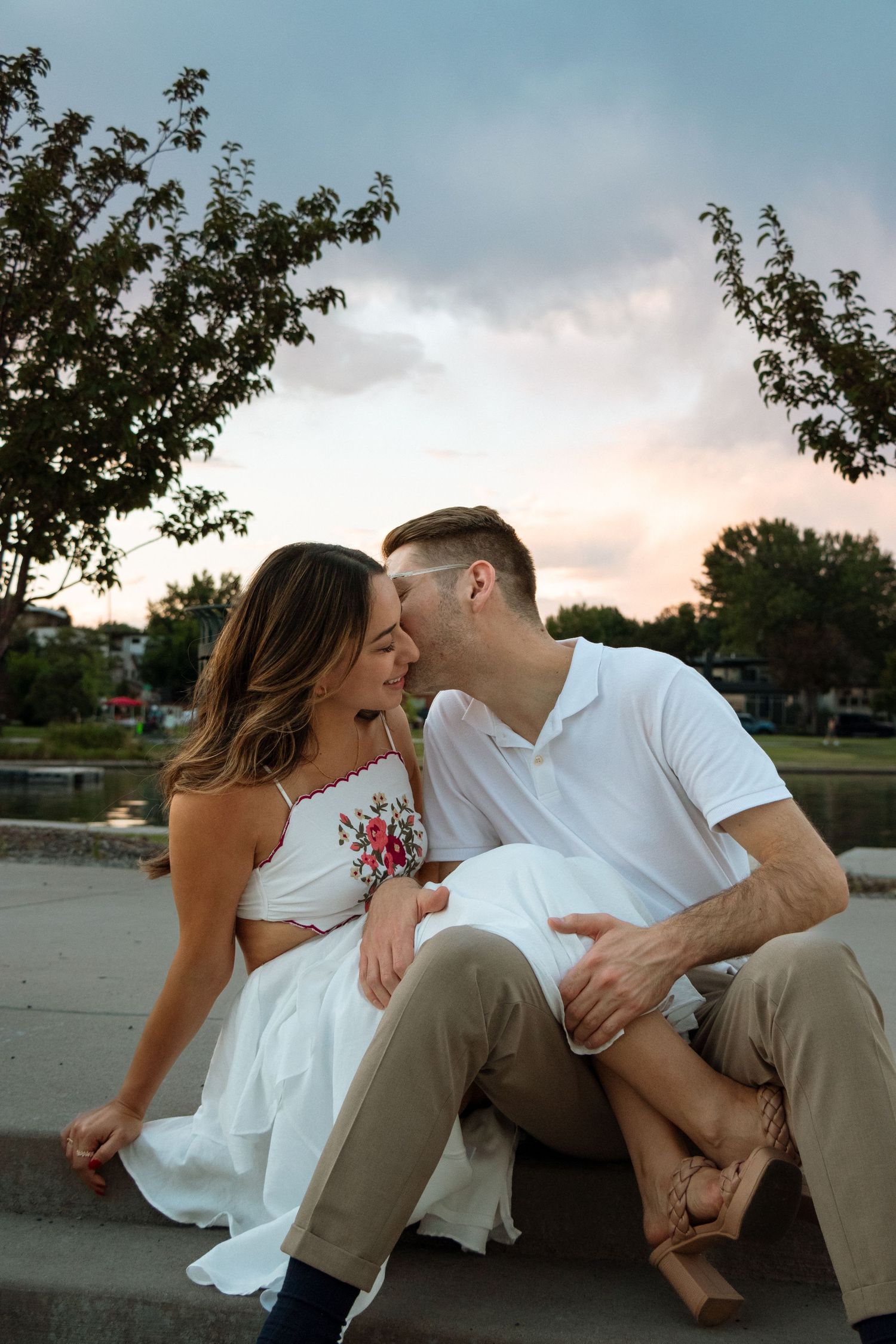 Romantic couple shares tender moments at sunset while sitting on concrete wall near a lake.