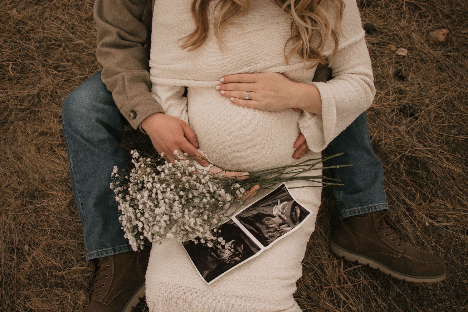 Pregnant couple sitting outdoors holding baby sonogram photos and white wildflowers, maternity announcement photo.