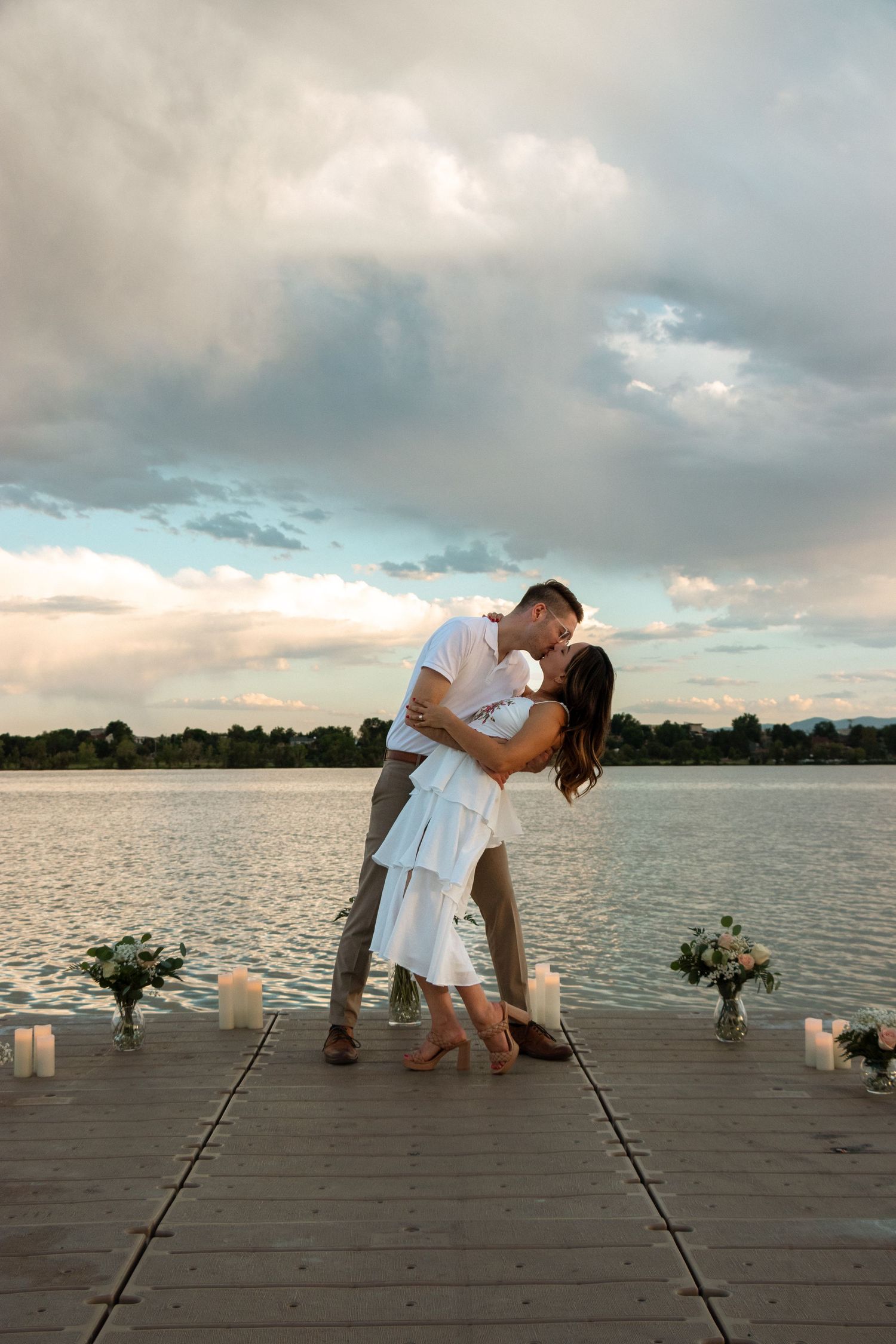 A couple in white embraces romantically on a wooden dock by a lake at sunset with candles and flowers.