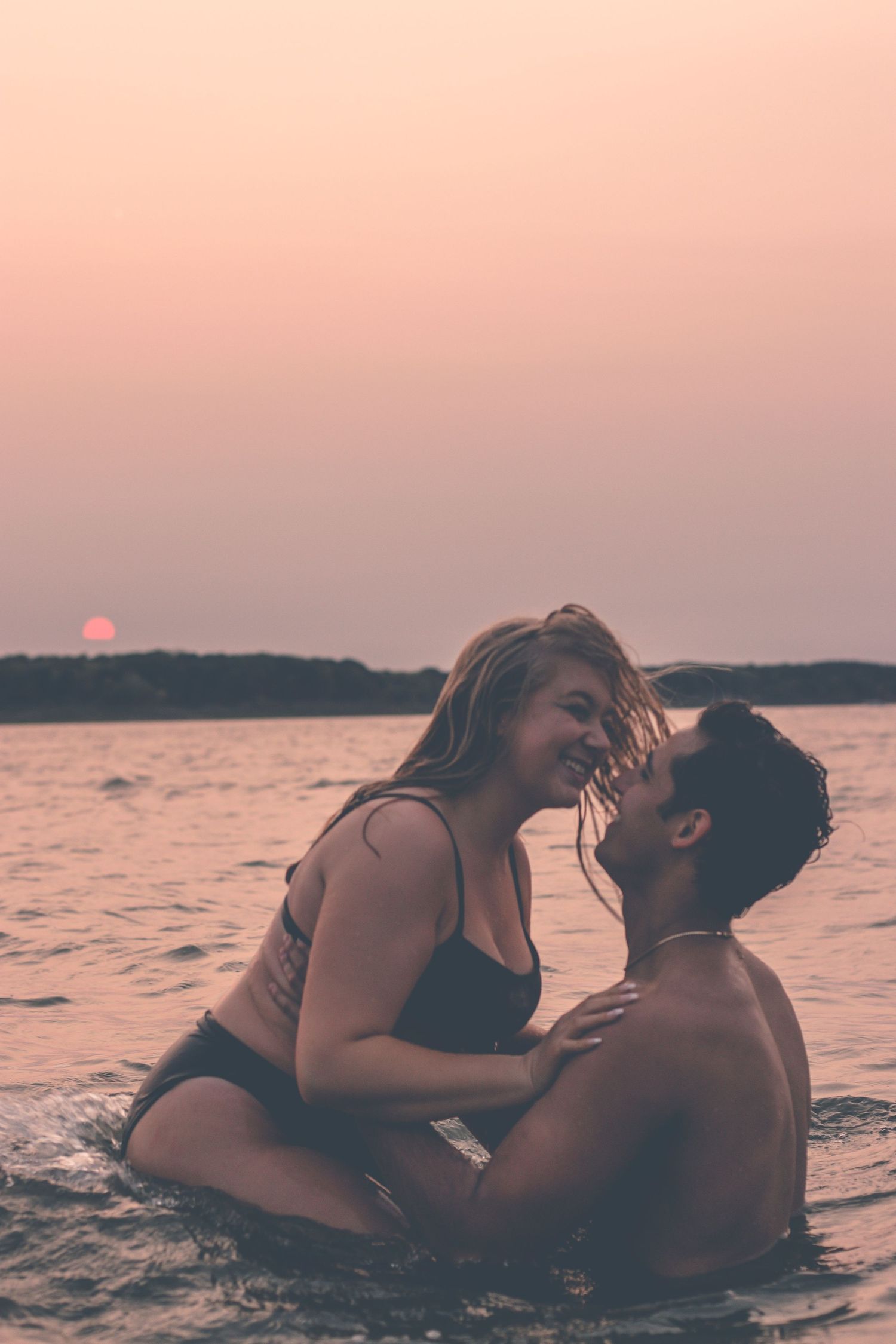 Couple embracing in lake water with pink sunset sky in background.