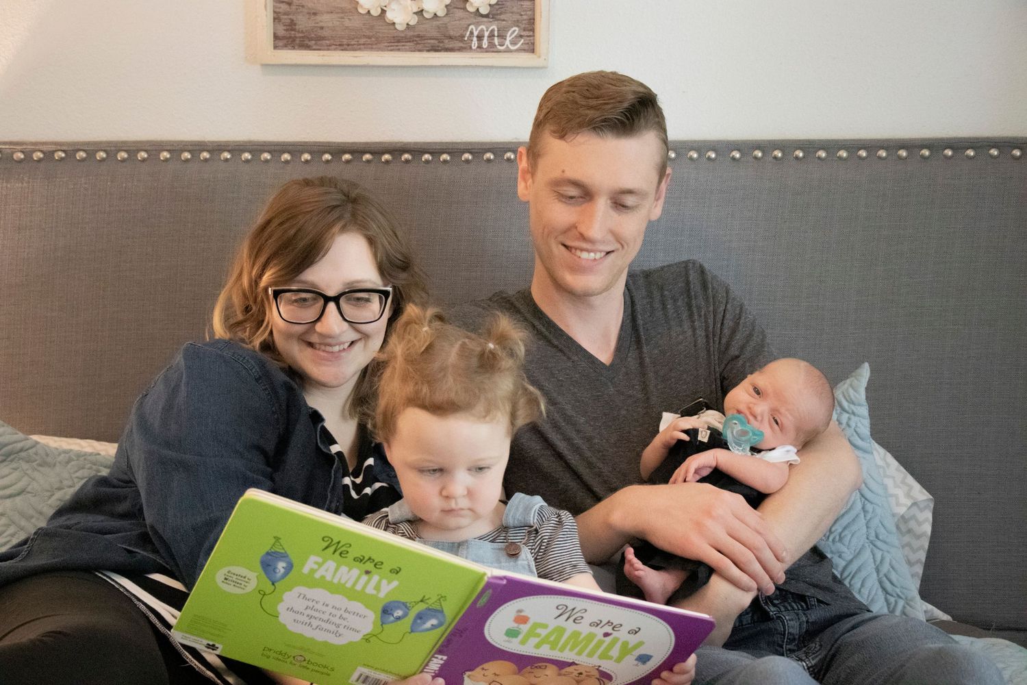A family reads children's books together on a gray couch with their newborn baby.
