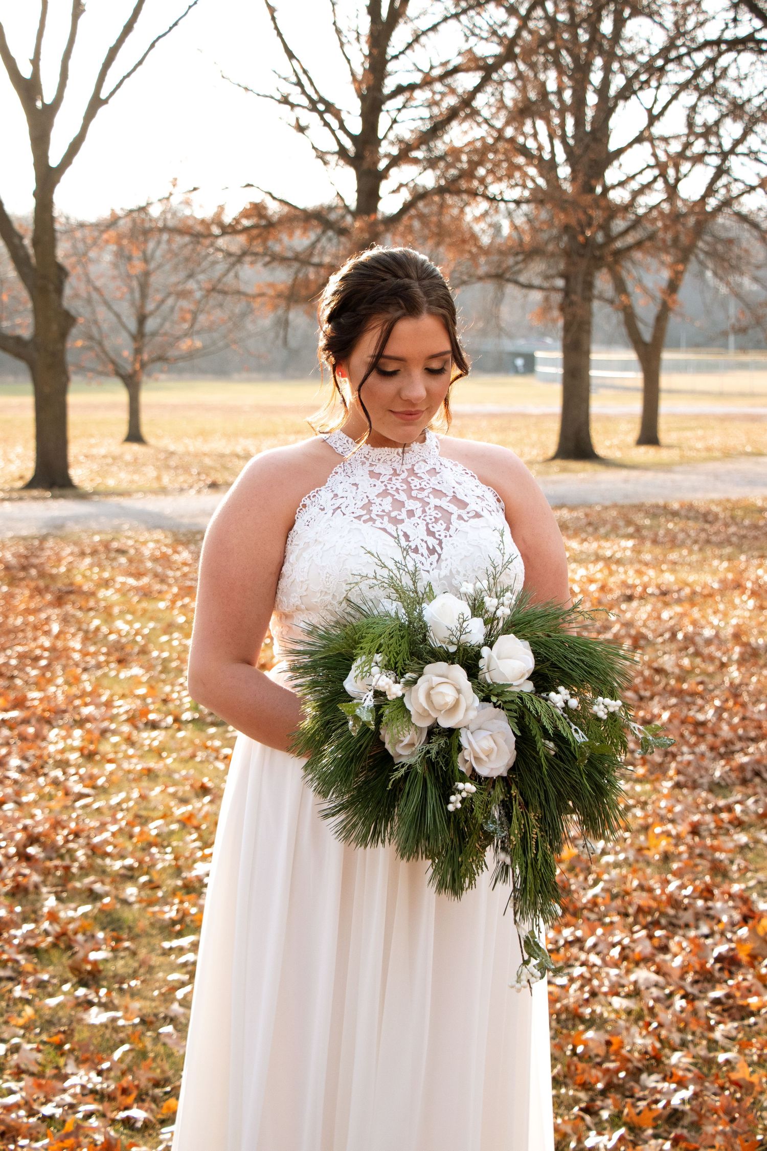 A white bridal bouquet with roses and evergreen branches against a backdrop of autumn leaves.