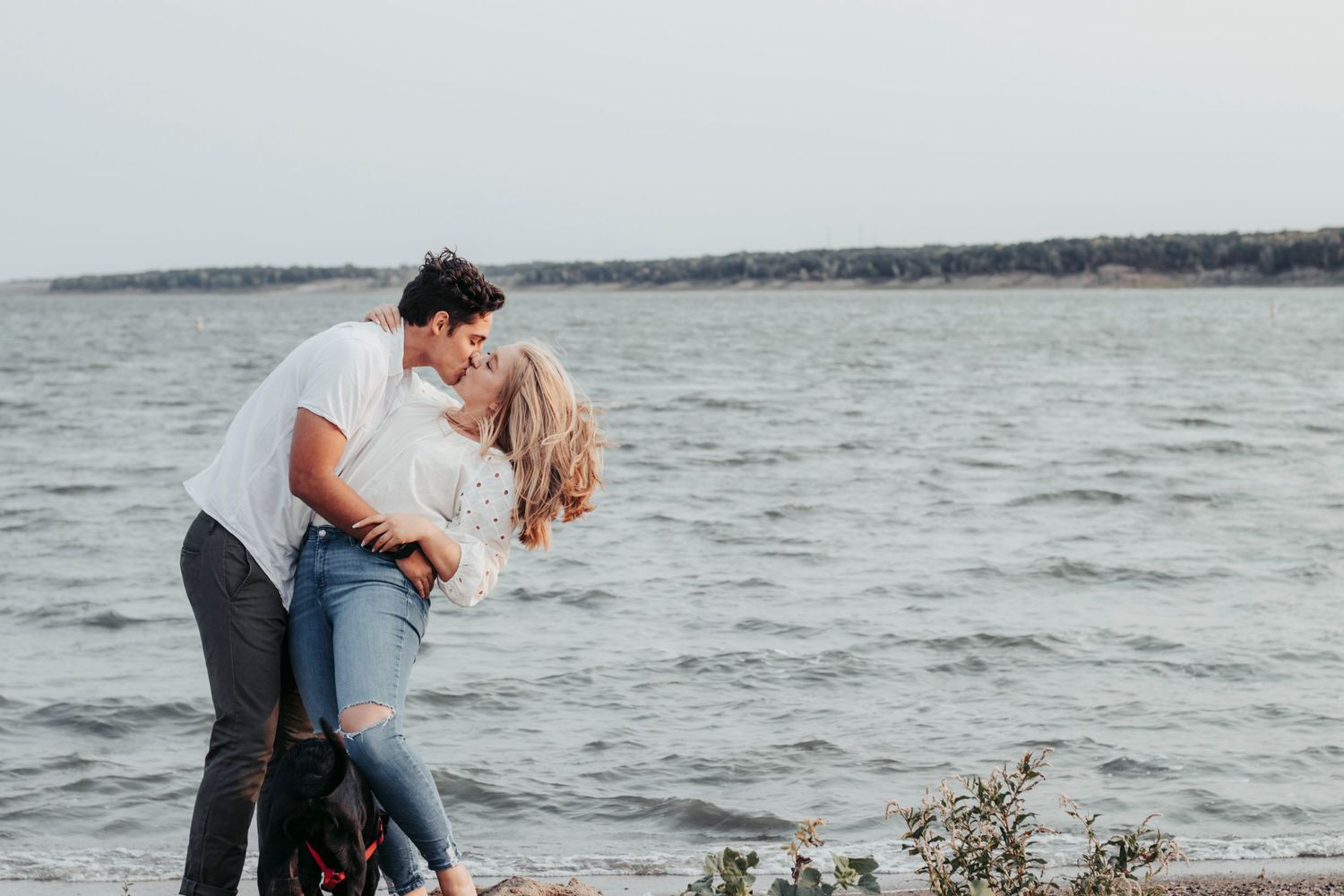 A couple embraces on the shoreline with water and trees in the background.