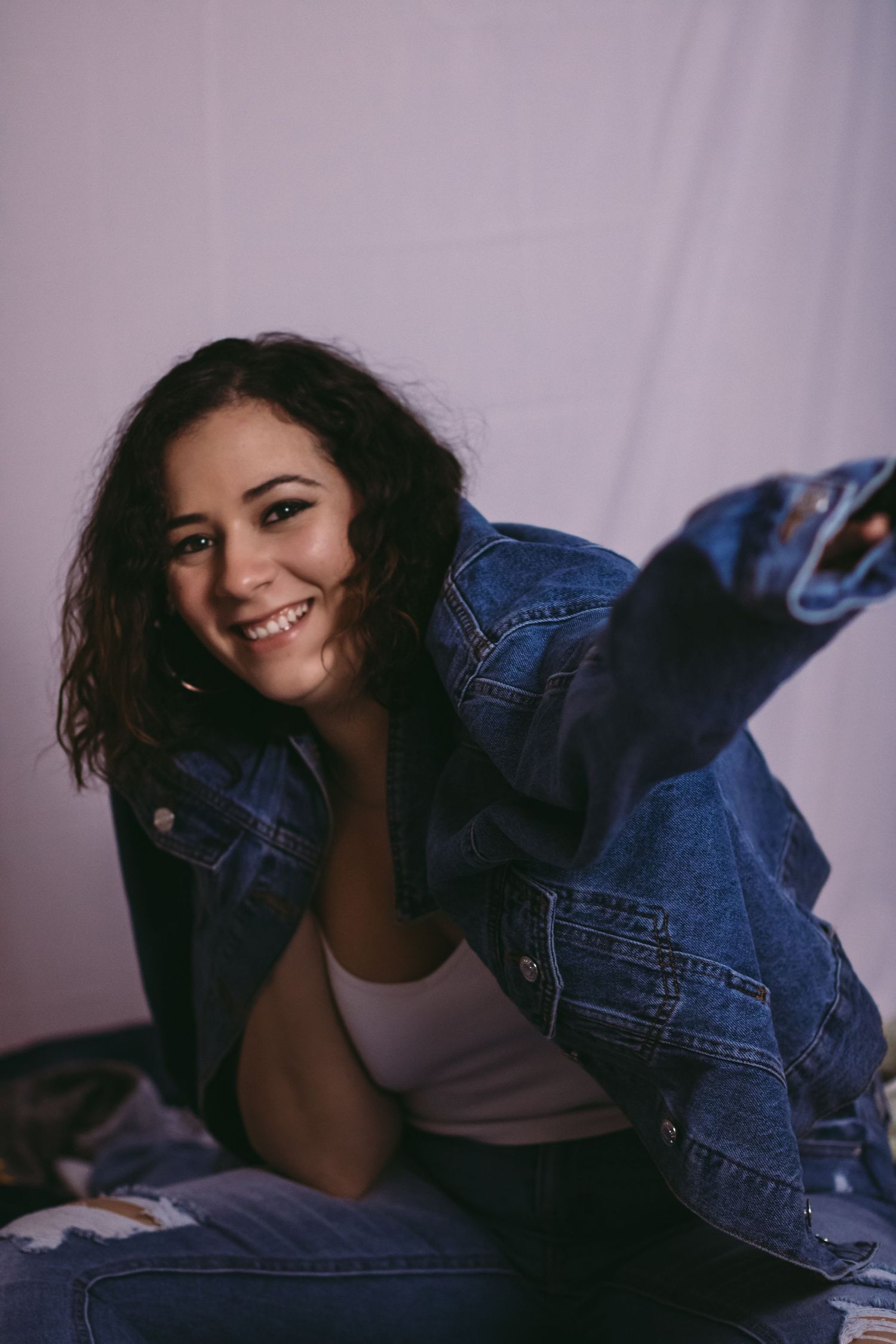 Model in denim jacket poses playfully against purple backdrop during casual fashion photoshoot.