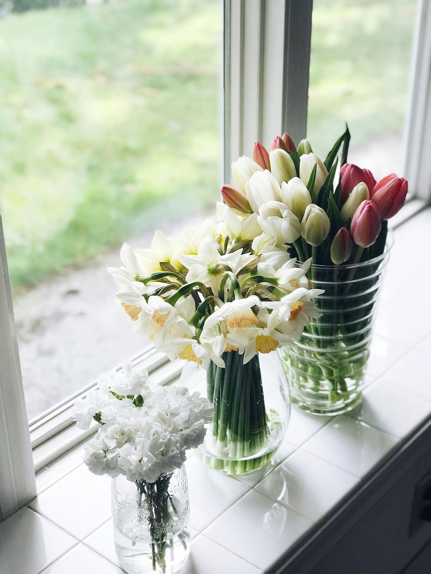 Fresh spring flowers including daffodils and tulips arranged in glass vases sit on a sunny windowsill.