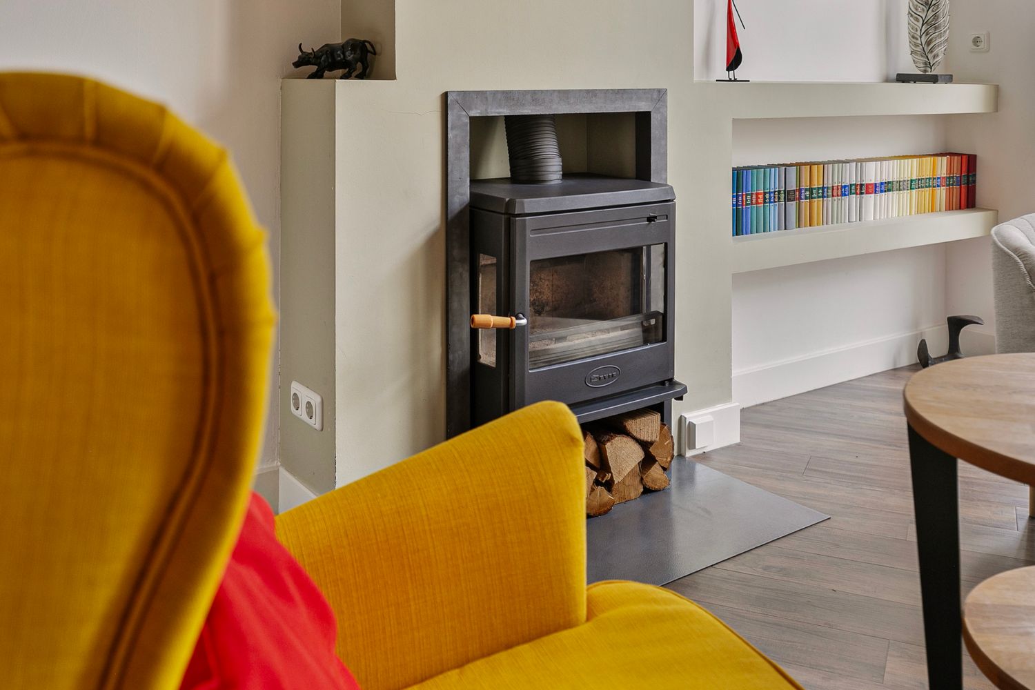 Modern living room interior with yellow accent chair and wood burning stove against white walls with built-in shelving.