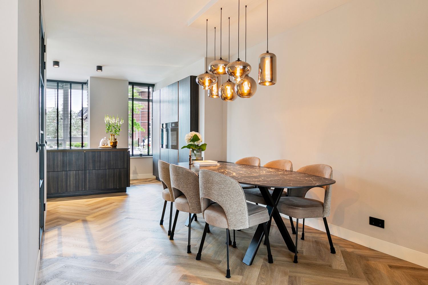 Modern dining area with gold pendant lights, upholstered chairs, and dark table on herringbone wood flooring.