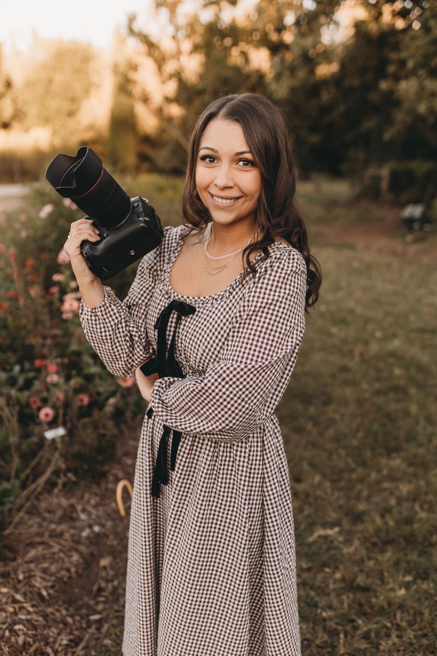 Professional photographer in gingham dress holding camera outdoors at sunset.