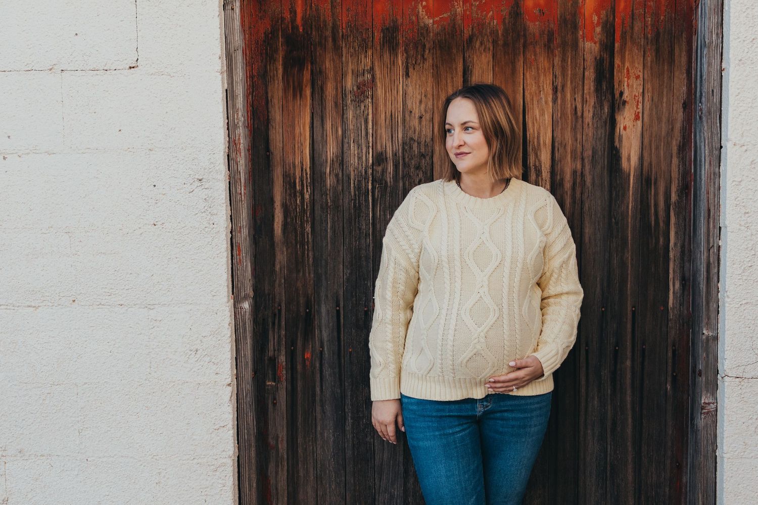 Woman in cream sweater and blue jeans stands against rustic wooden wall while holding smartphone.