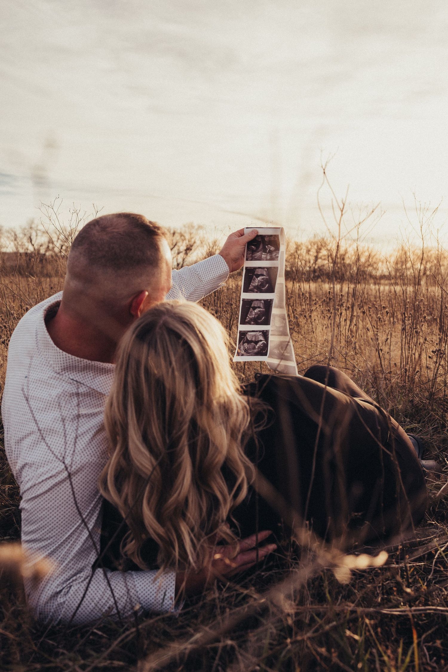 Couple lying in autumn field holding ultrasound photos at sunset.