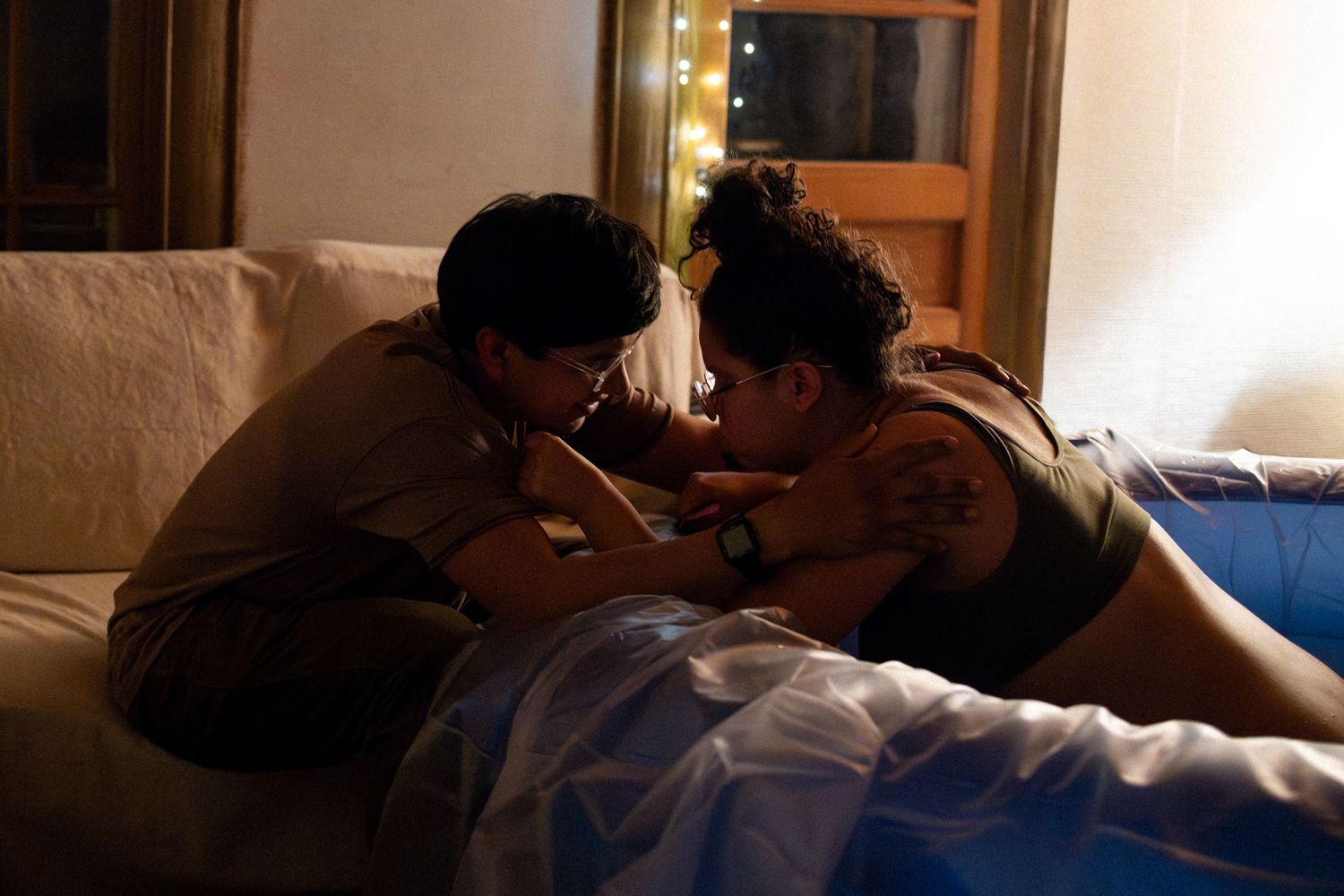 Two people share an intimate moment lying together on a bed in a warmly lit room with fairy lights.