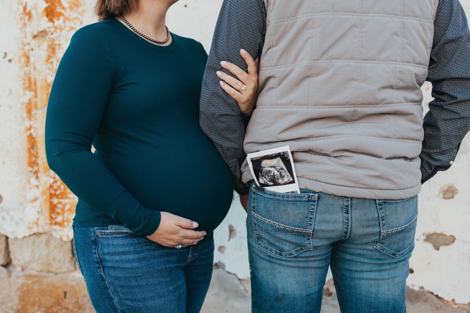 A couple wearing blue jeans and casual tops embrace while standing against a rustic white wall.