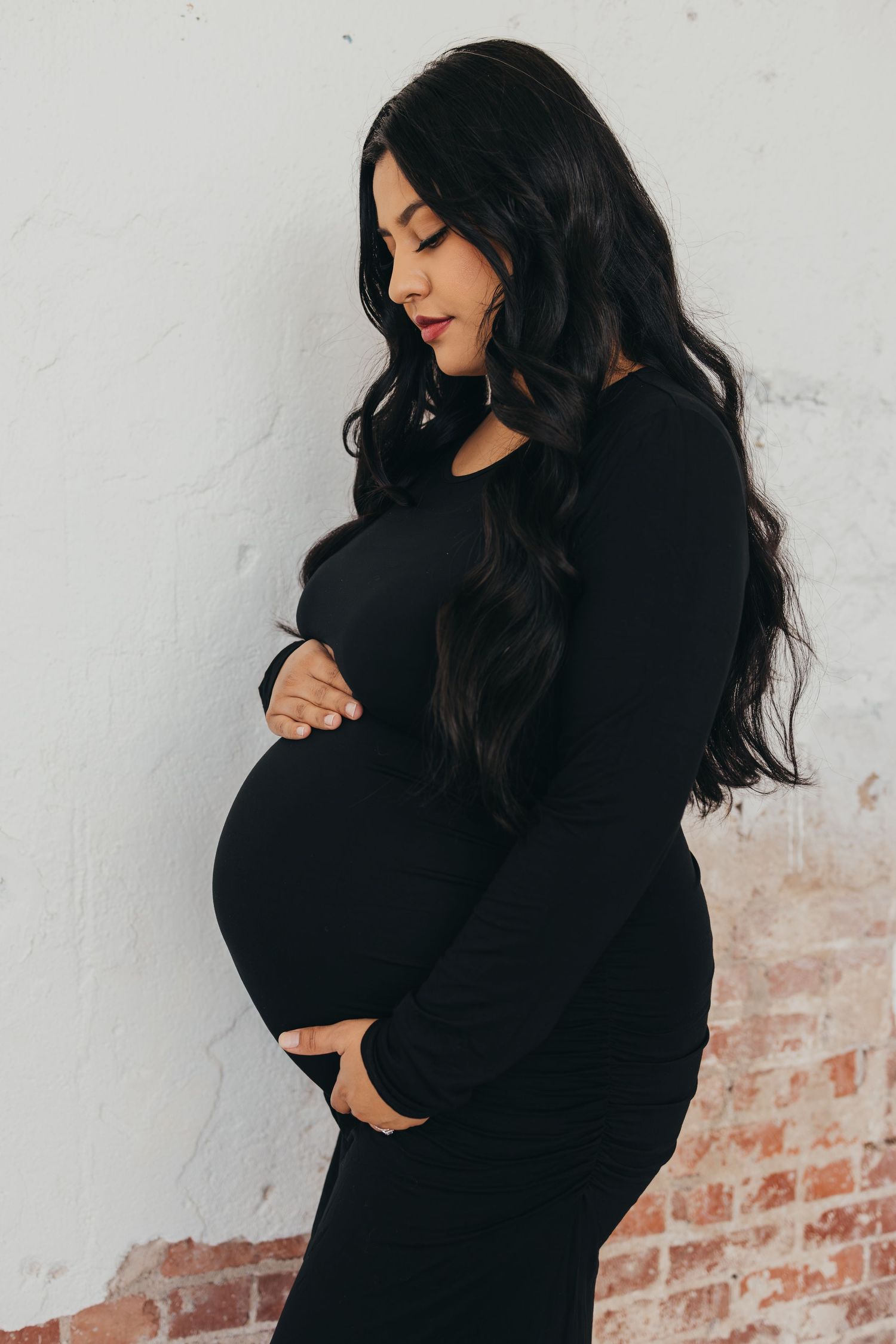 Artistic black maternity dress photos against white brick wall.