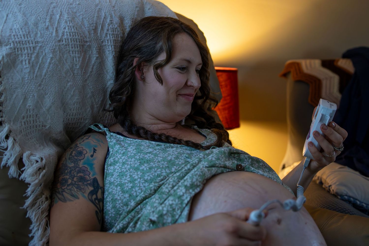 A pregnant person in a floral dress looks at their phone while relaxing in bed with warm lighting.