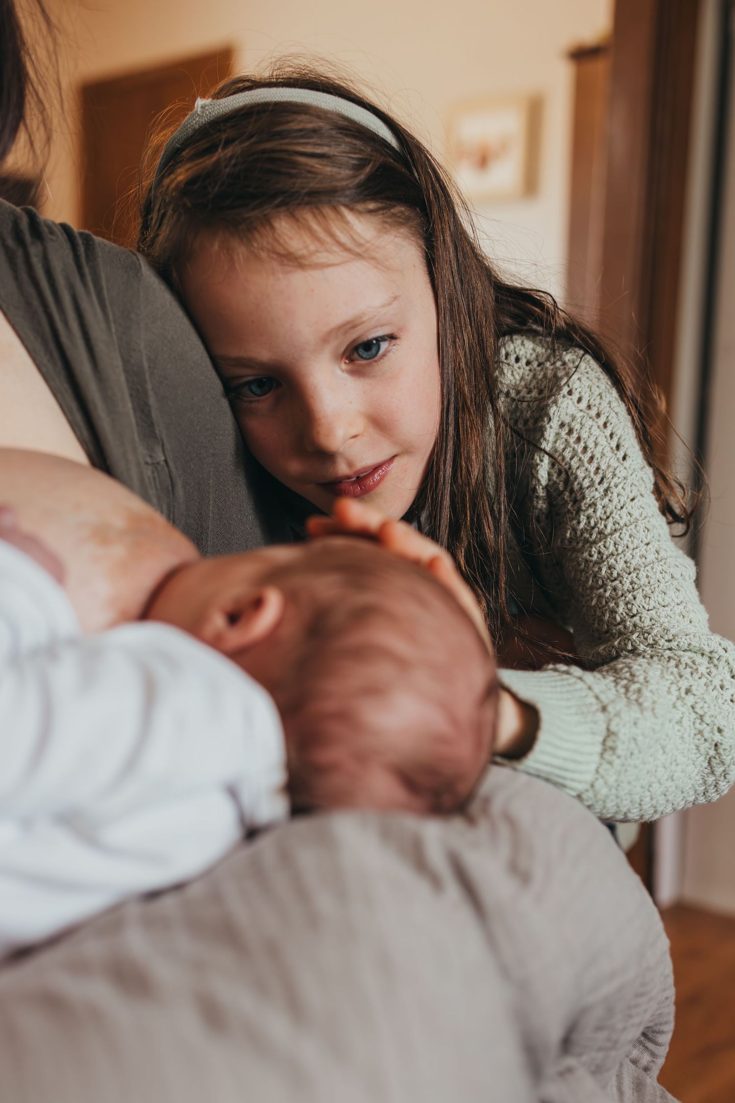 A child in a knit sweater leans over to kiss a sleeping baby in a cozy indoor setting.