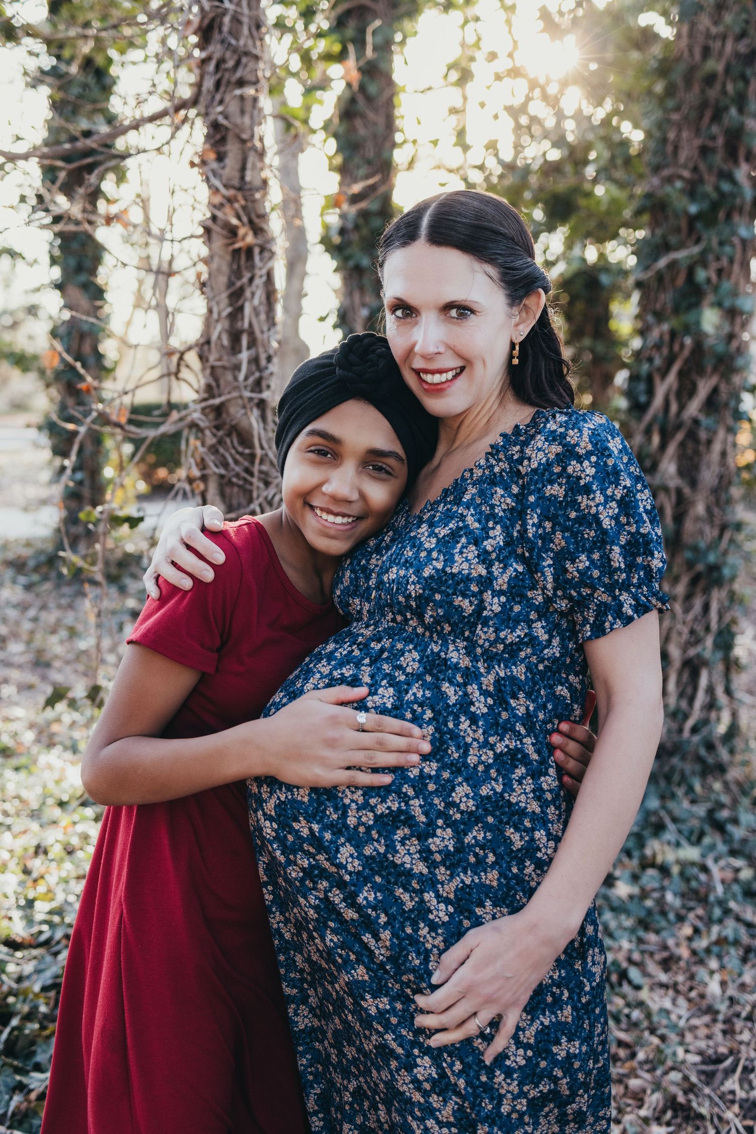 Outdoor maternity photoshoot with two people embracing against a sunlit wooded background.