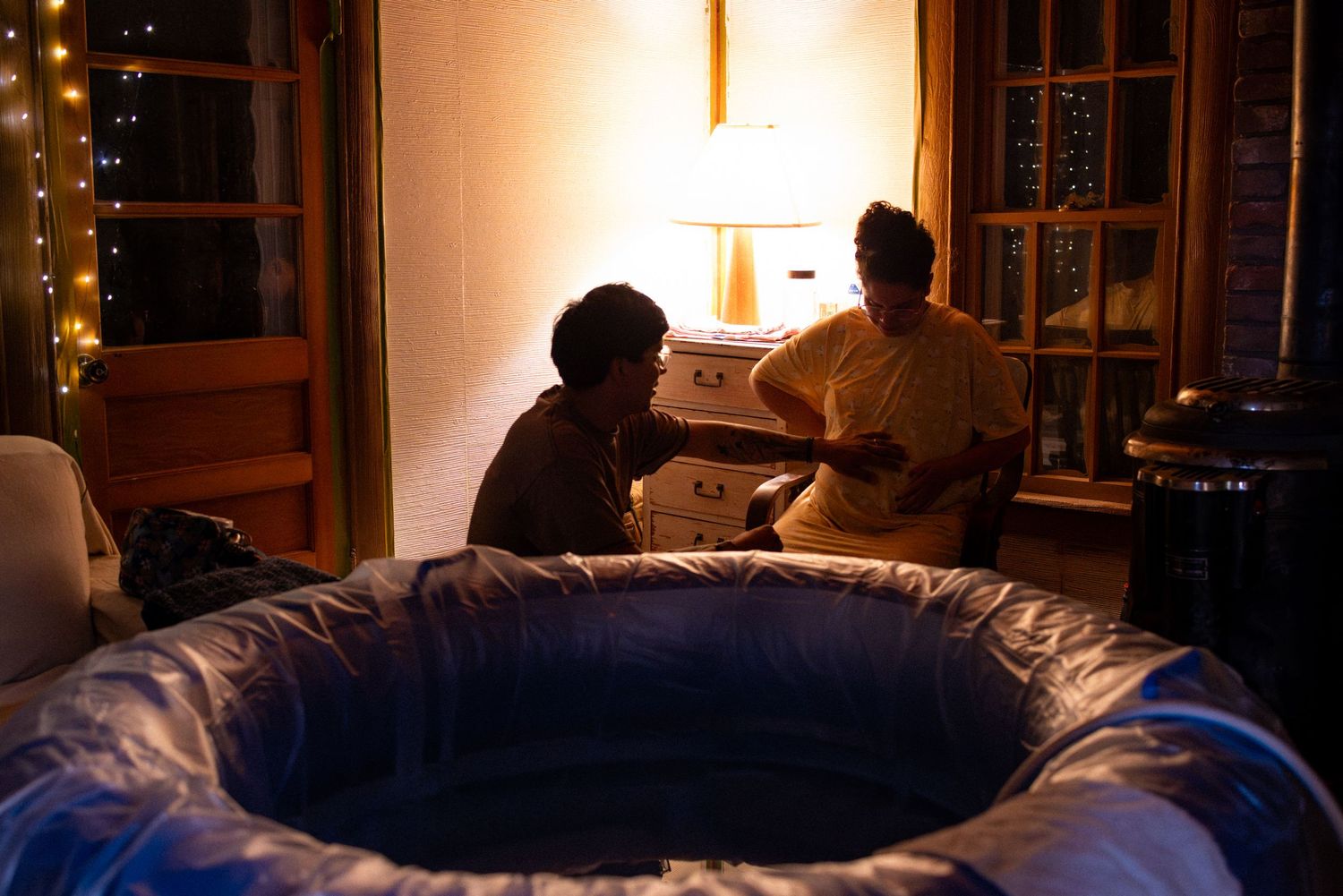 Two people sit beside an inflatable pool indoors at night, with warm lamp light and string lights in background.
