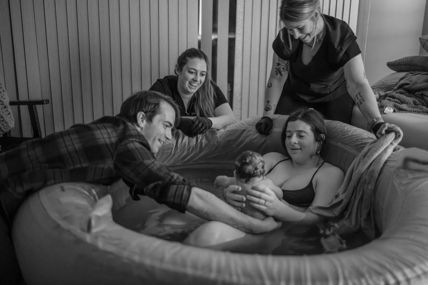 A black and white photograph of a home birth scene with support people gathered around a birthing pool.